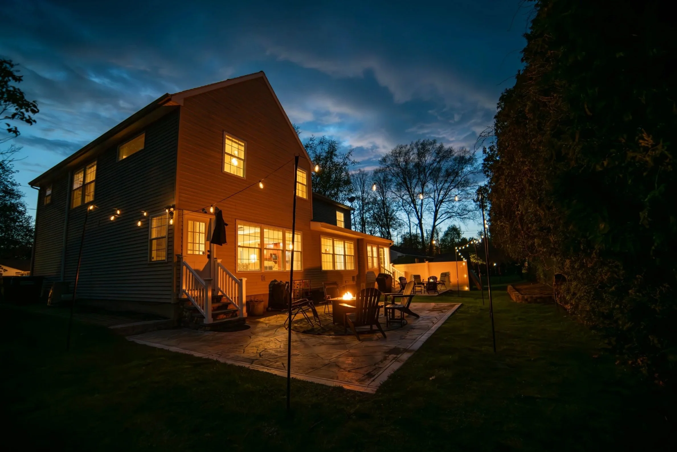 A two-story house with illuminated windows at dusk, surrounded by string lights hanging outside, a patio with chairs, a fire pit, and a lawn with trees in the background.