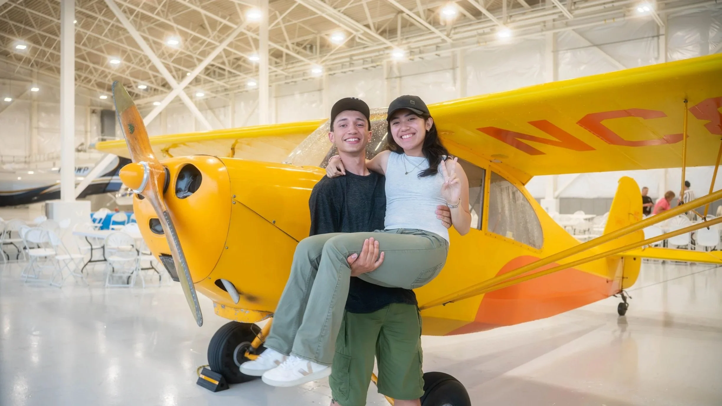A young man holding a young woman in front of a yellow airplane inside an aircraft hangar.