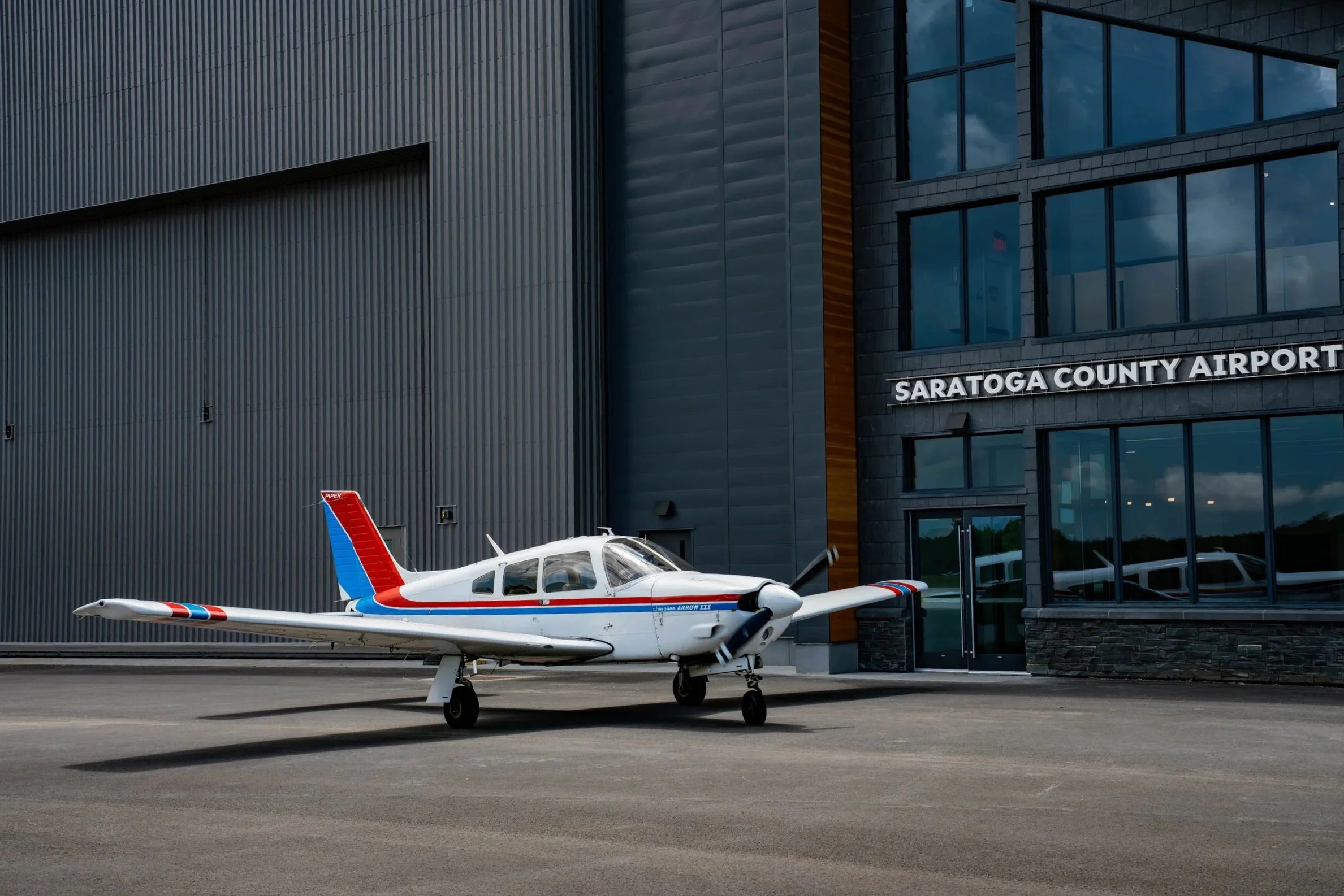 A small airplane parked outside Saratoga County Airport building with modern glass and metal exterior.