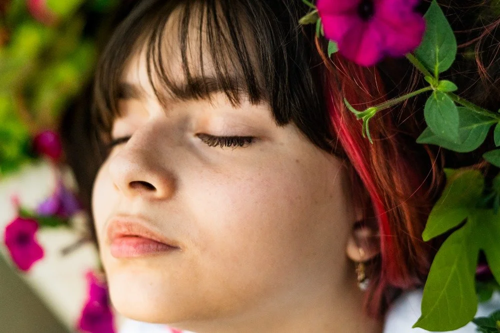 A young woman with dark hair and a red streak, closed eyes, surrounded by pink and purple flowers and green leaves.