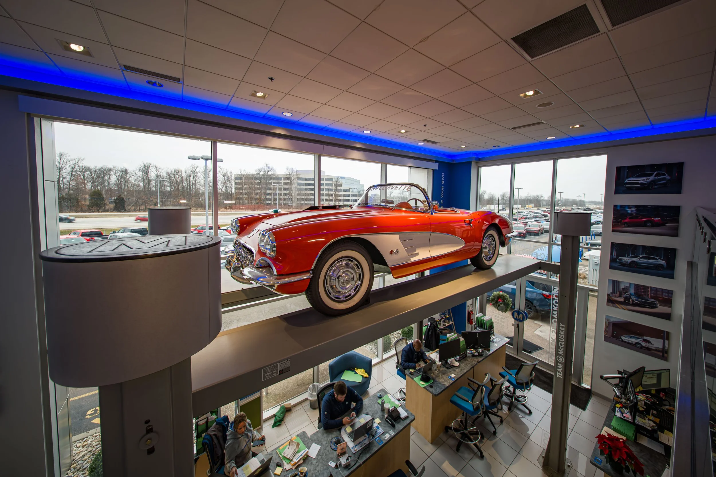 A red, vintage Chevrolet Corvette car displayed on a raised platform inside a car dealership showroom, with large windows showing parking lot and trees outside.