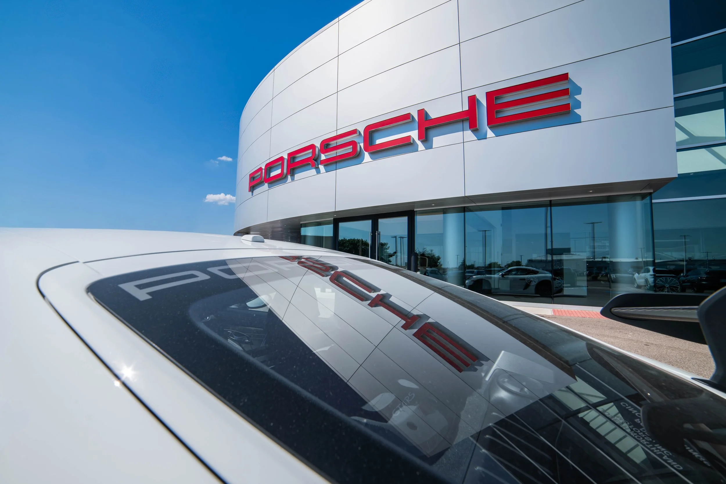 A white car parked outside a Porsche dealership with the Porsche logo on the building in the background.