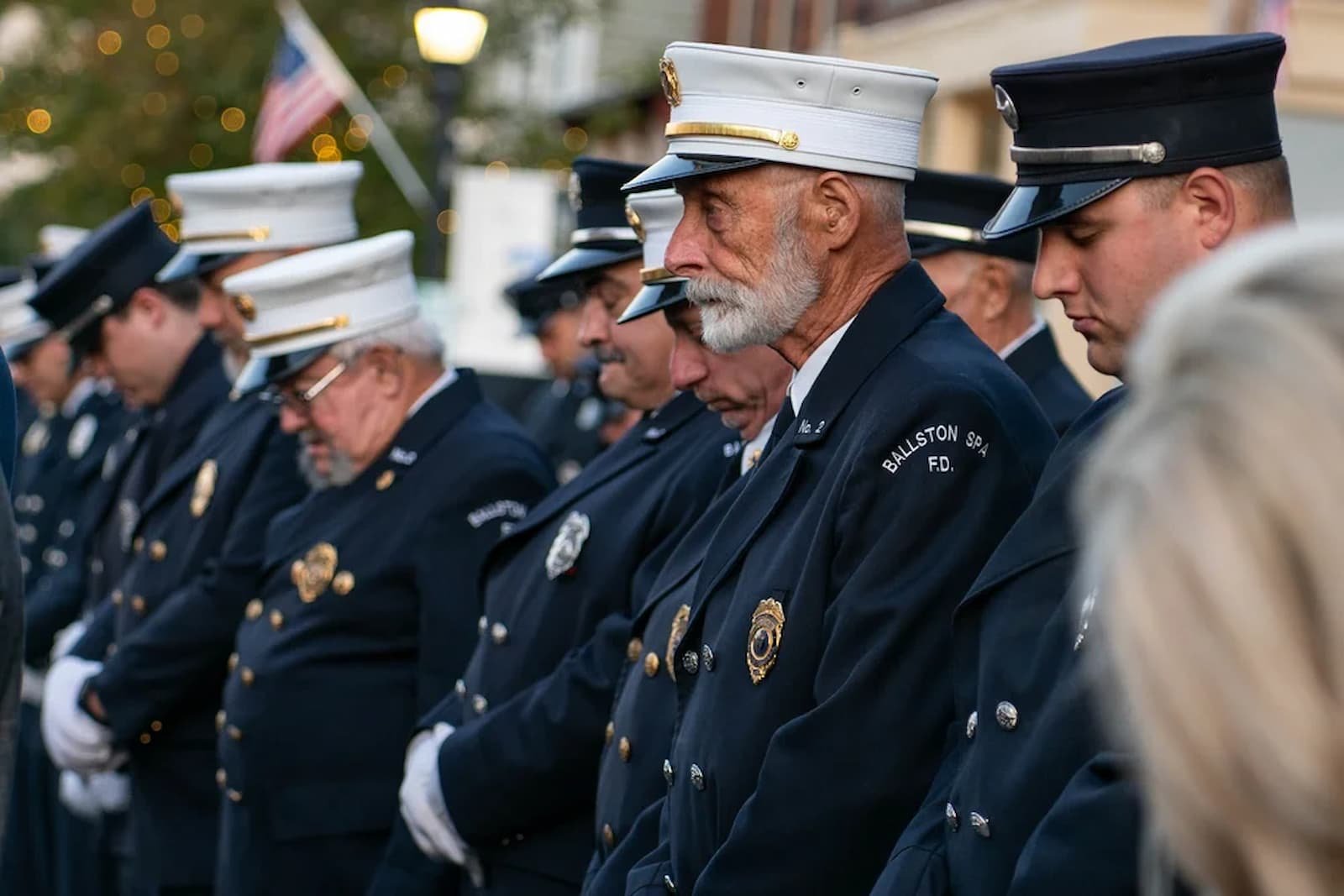 A line of firefighters in dress uniforms and hats bowing their heads during a memorial or ceremonial event.