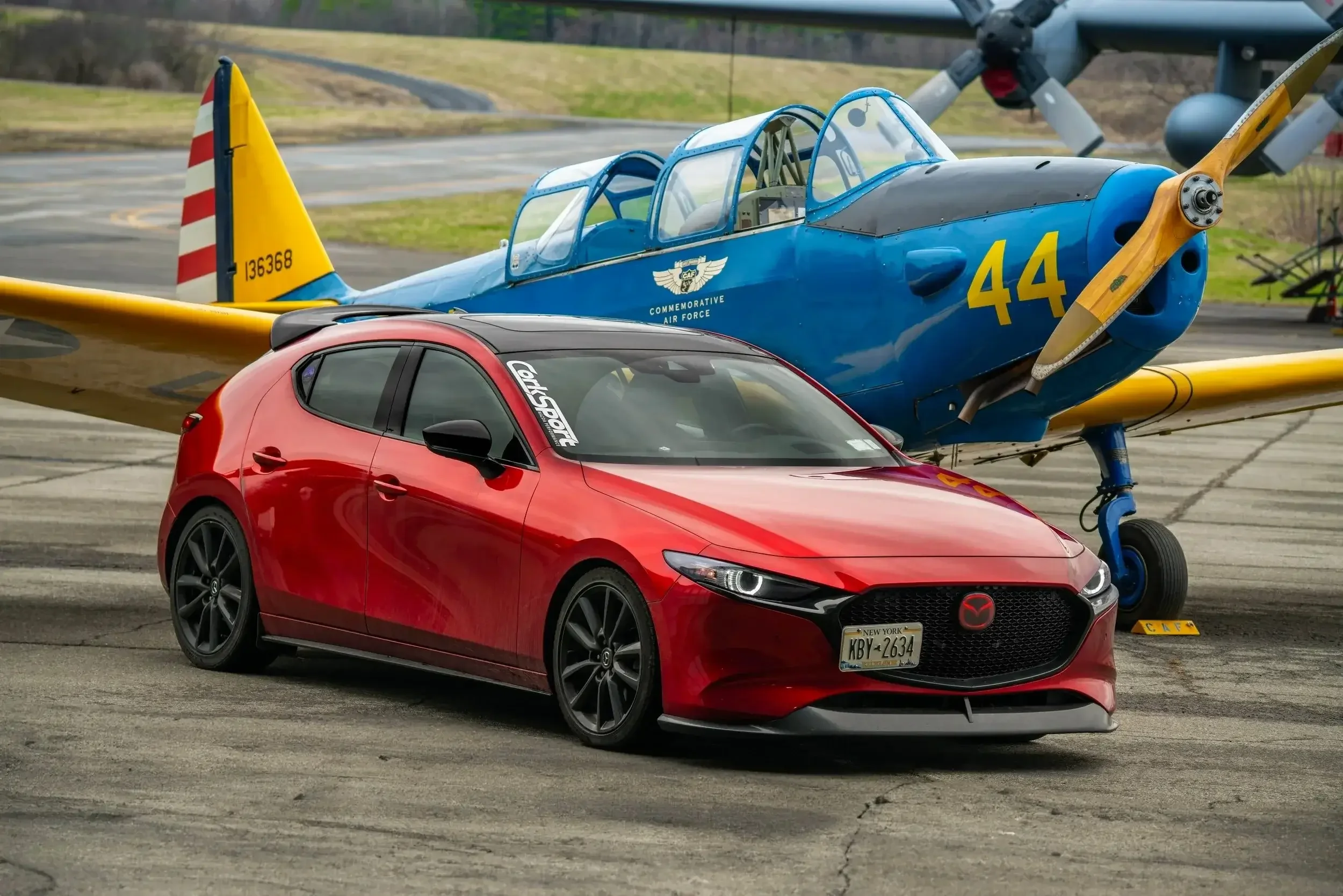 A red Mazda hatchback car parked on an airfield in front of a vintage blue and yellow airplane.