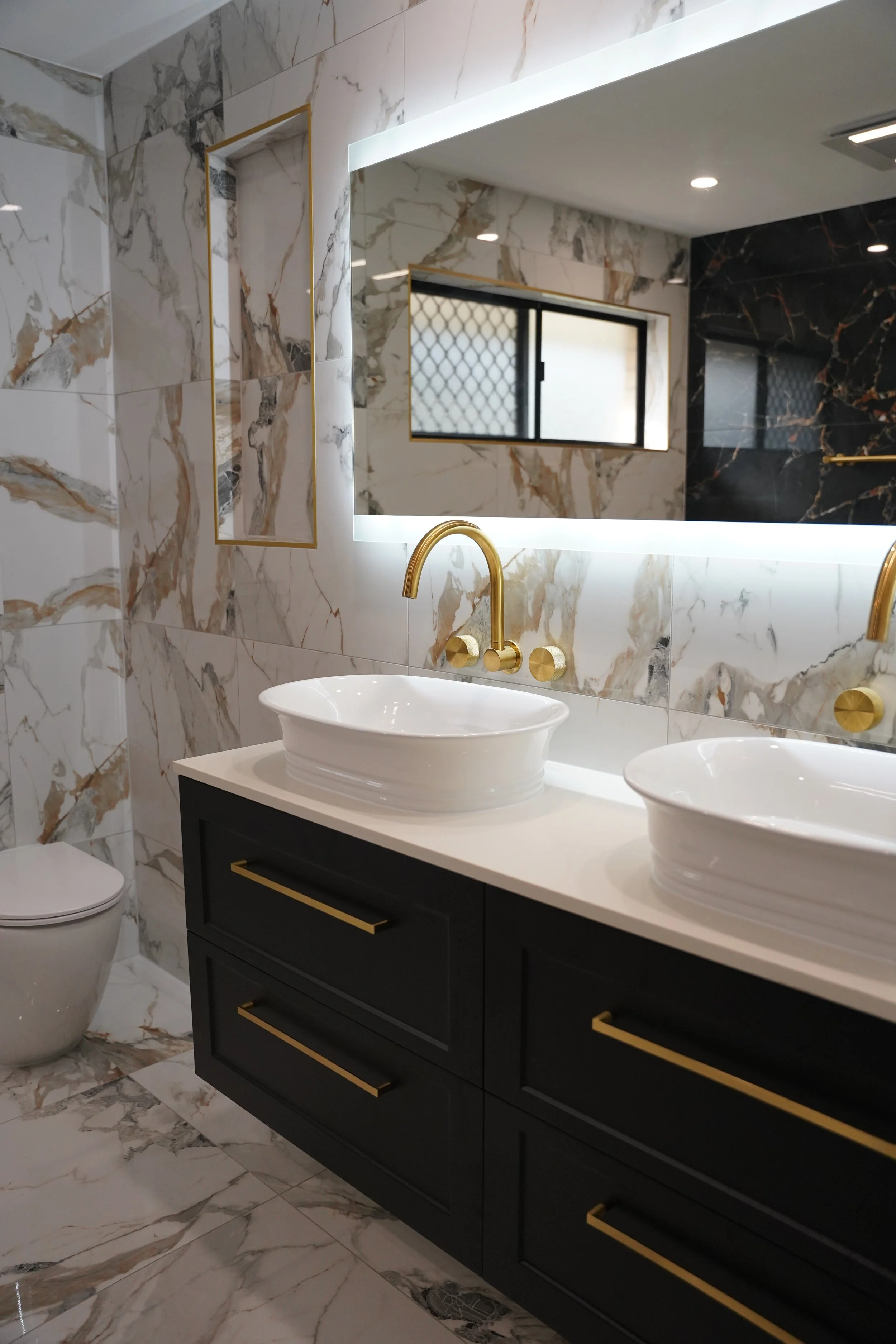 Modern bathroom with marble tiles on walls and floor, dual white vessel sinks on a black vanity with gold handles, a large backlit mirror, gold wall-mounted faucets, a window with security mesh, and a white toilet.