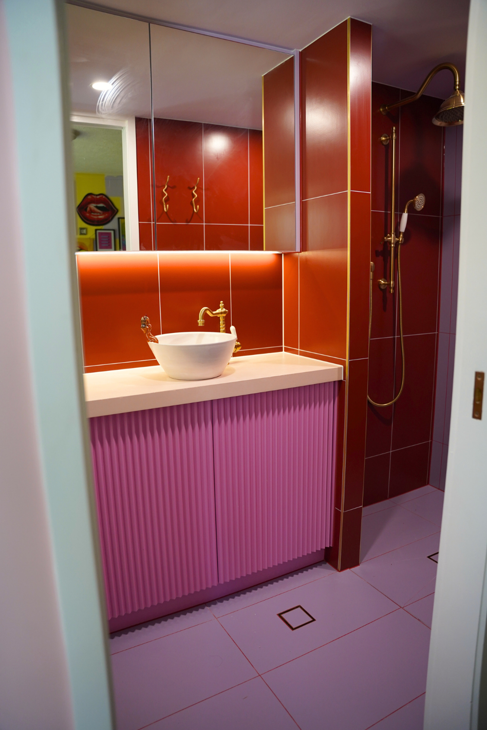 A modern bathroom with pink and red tiles, a white vessel sink with a gold faucet, mirrored cabinets, and a walk-in shower with gold fixtures.