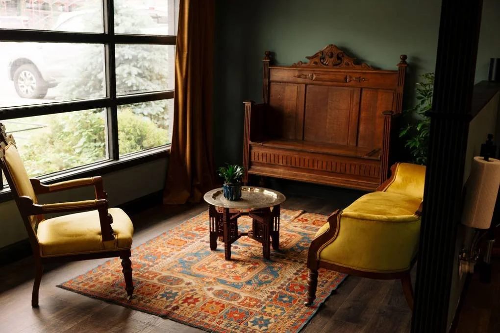Living room corner with vintage yellow upholstered armchair and small sofa, wooden hall tree, round wooden coffee table with potted plant, colorful patterned rug, large window with brown curtains, and a green wall with a decorative plant.