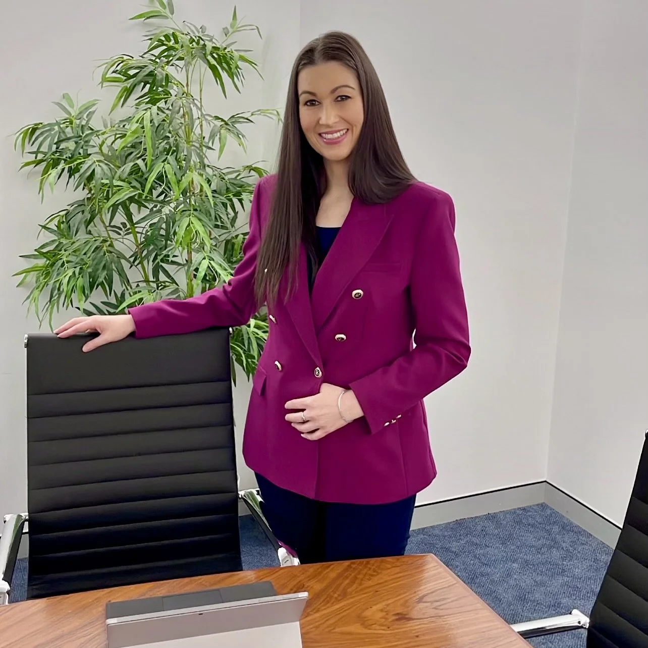 A woman with long brown hair wearing a bright pink blazer standing in a conference room, resting her hand on the back of a black office chair, with a large green plant in the background.