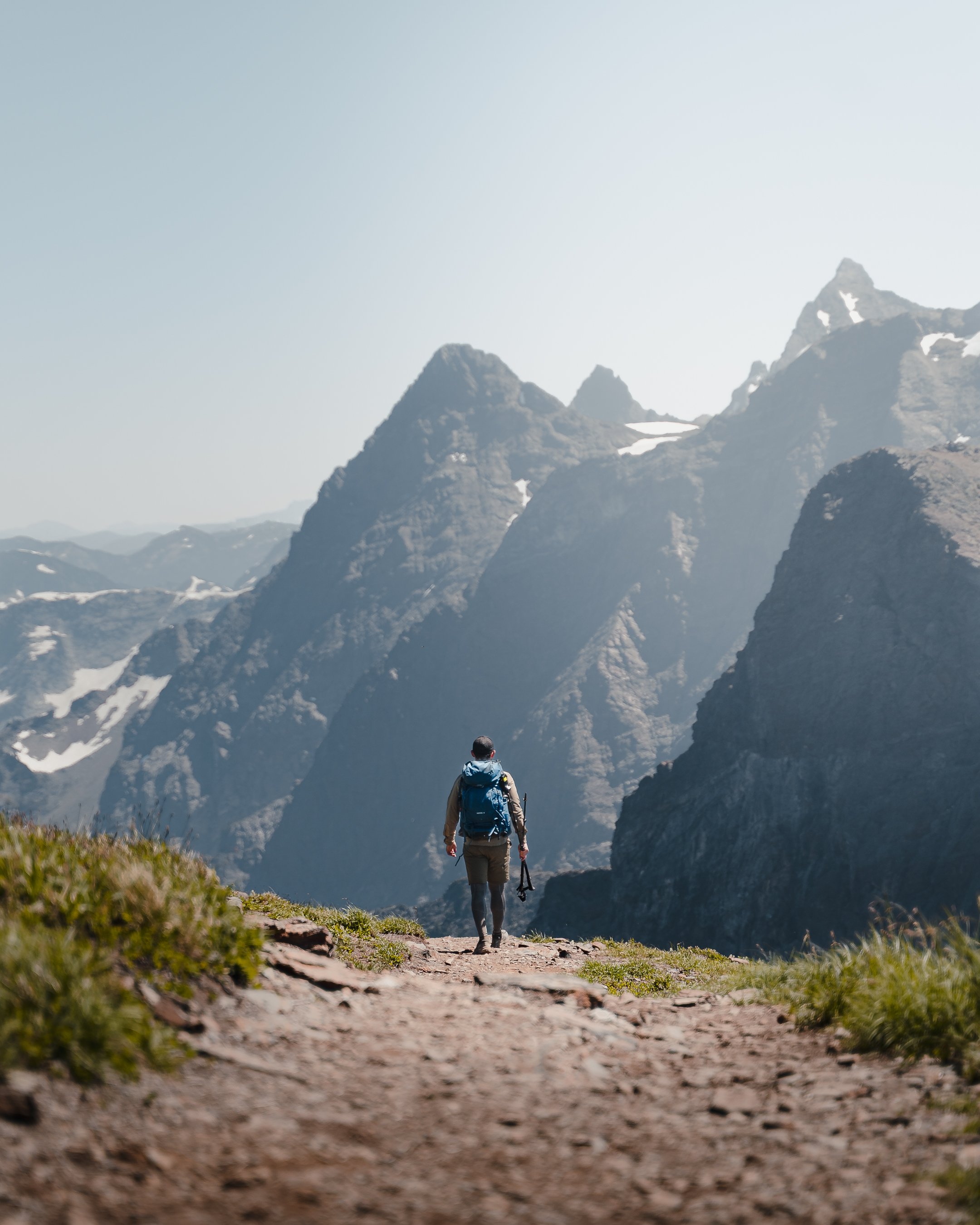 Hiker with backpack walking on a mountain trail overlooking rugged peaks.