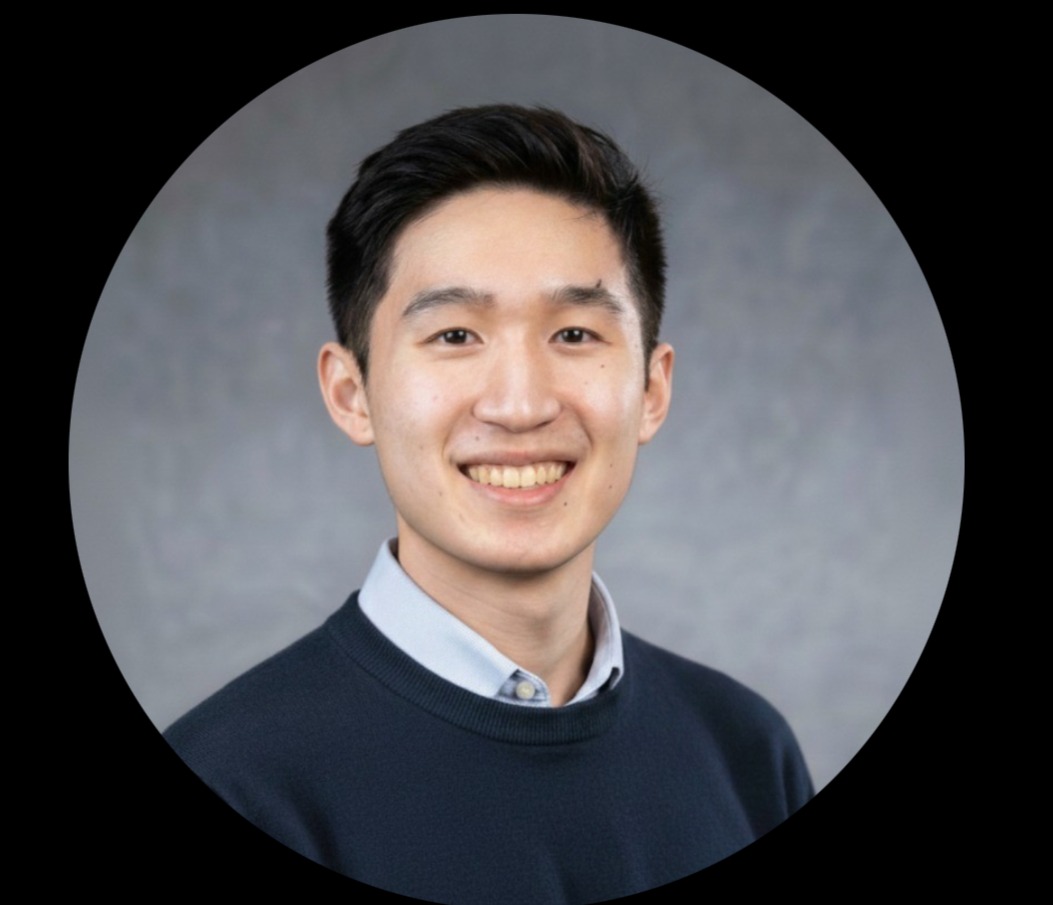 A young man with black hair, smiling, wearing a dark sweater over a collared shirt, with a gray background.