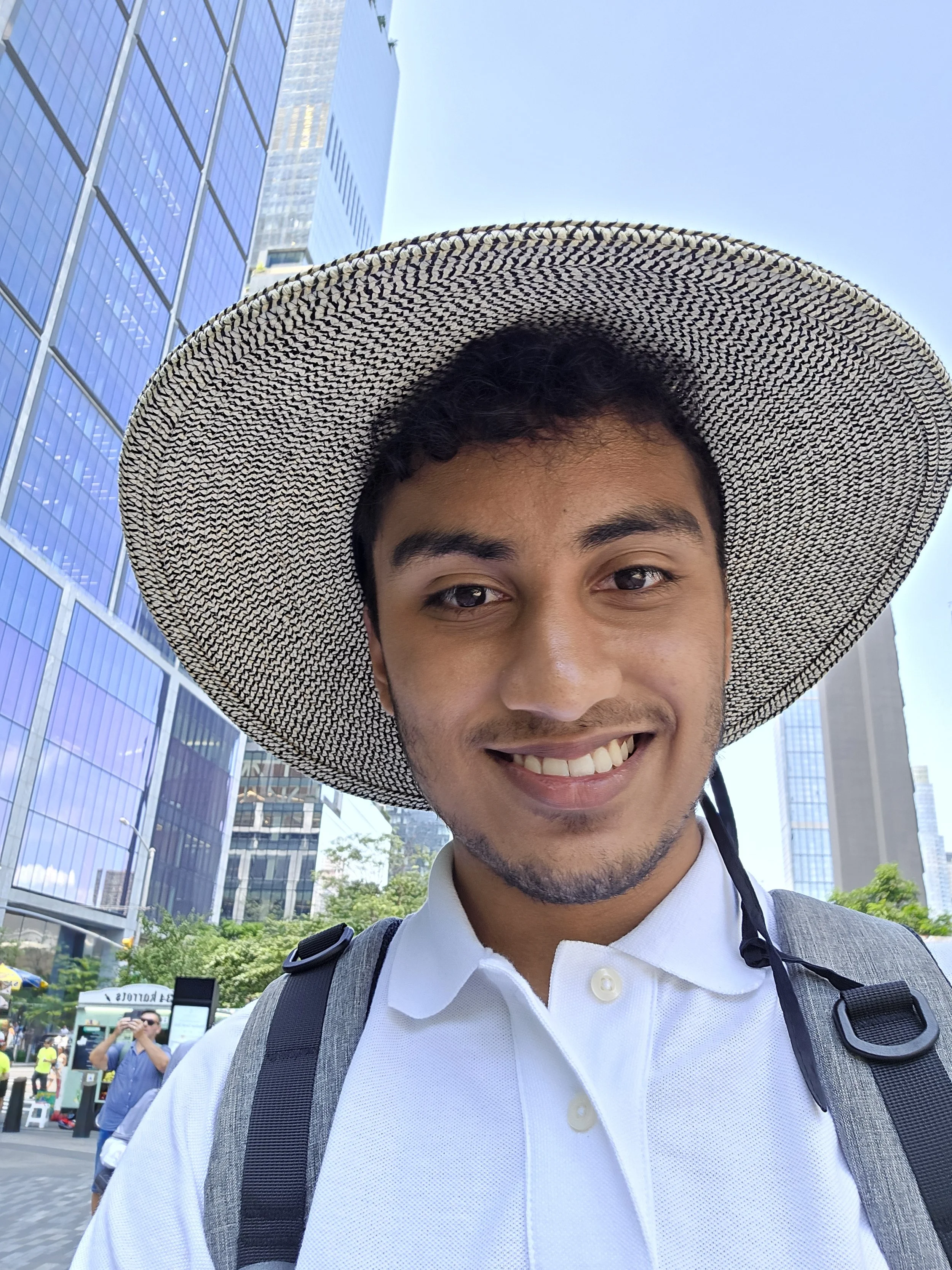 A young man smiling outdoors in a city setting, wearing a wide-brimmed hat, a white polo shirt, and a backpack.