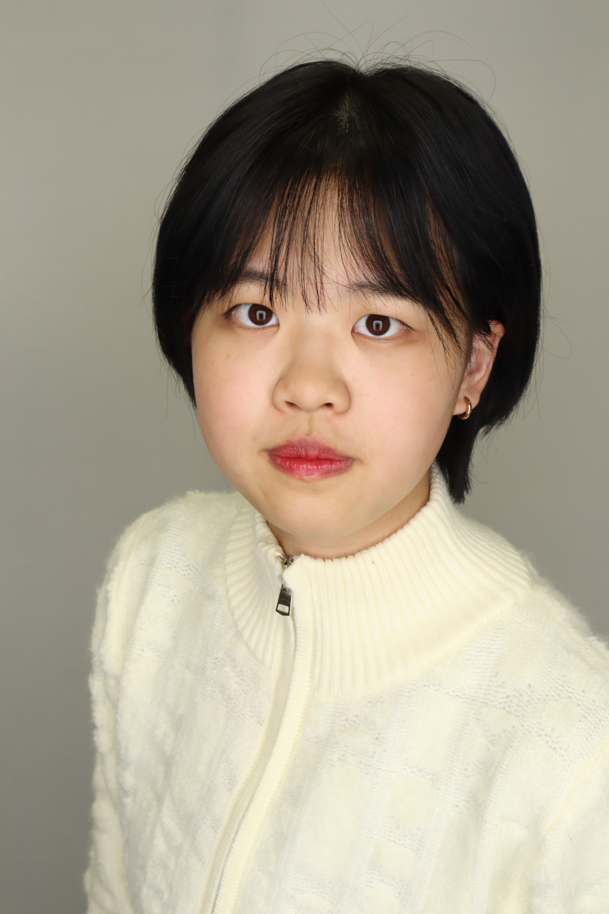 Close-up portrait of a young woman with short black hair, wearing a cream-colored zip-up sweater, standing against a plain background.