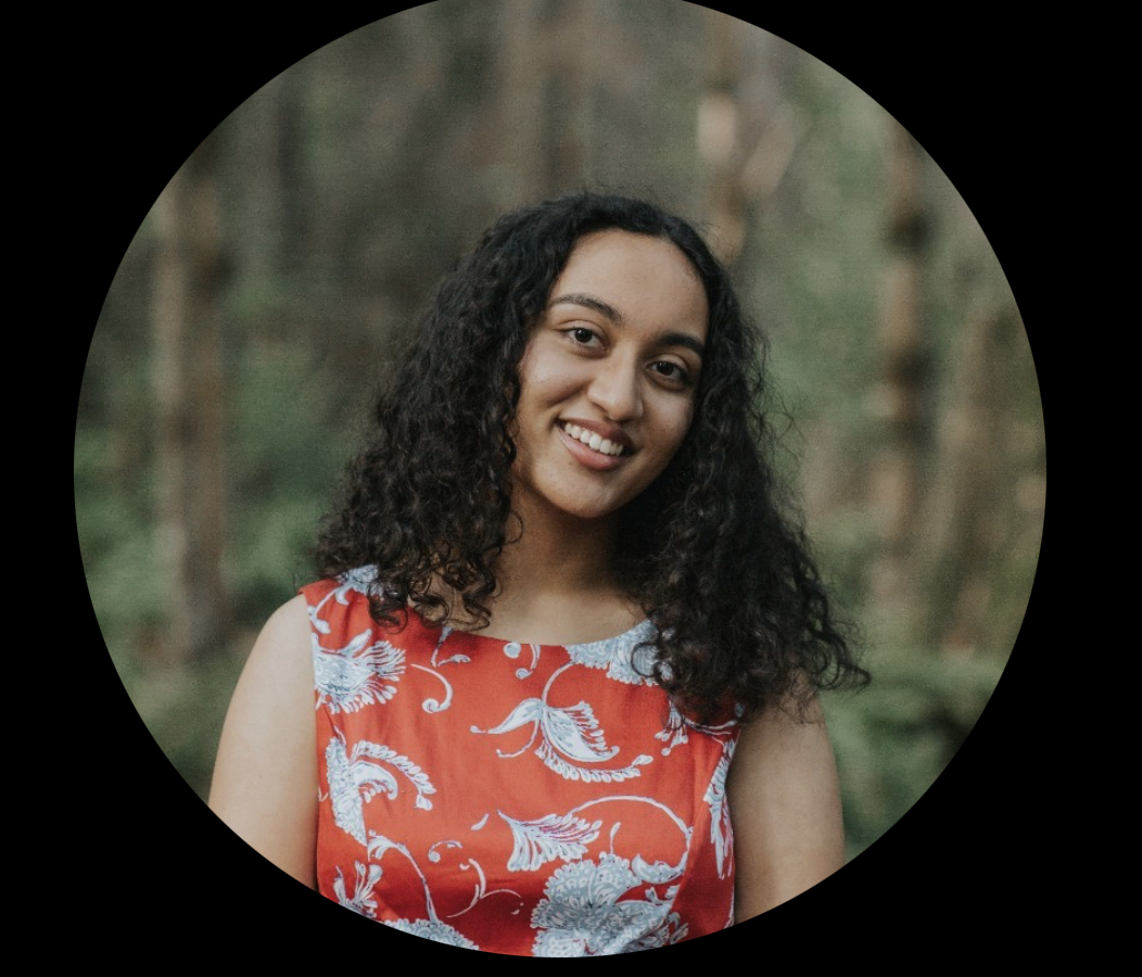 A woman with dark curly hair smiling outdoors, wearing a sleeveless red dress with white floral patterns, in a forest setting.
