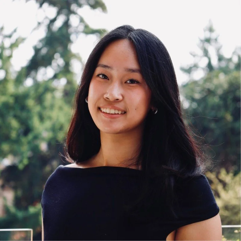 A young woman with shoulder-length black hair smiling outdoors with trees in the background.
