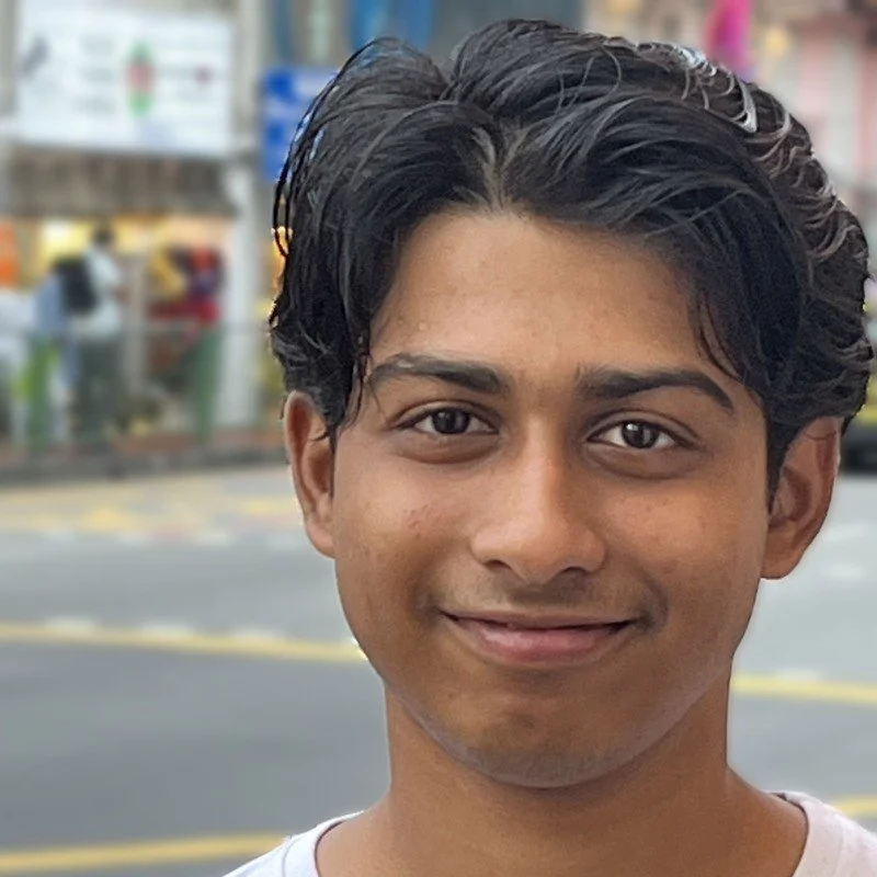 A young man with dark, wavy hair and a light smile, standing outdoors in an urban area with a blurred storefront and street background.