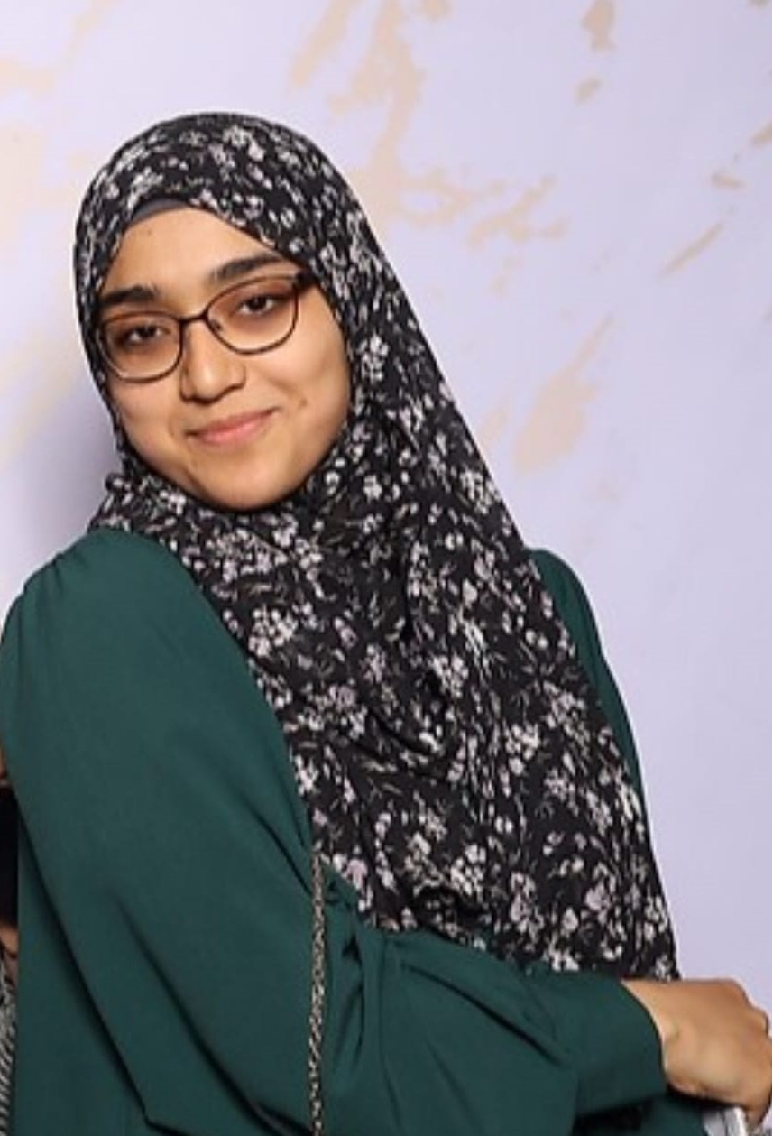 A young woman with glasses wearing a black and white floral hijab and a dark green top posing with a smile against a light-colored background with faint abstract markings.