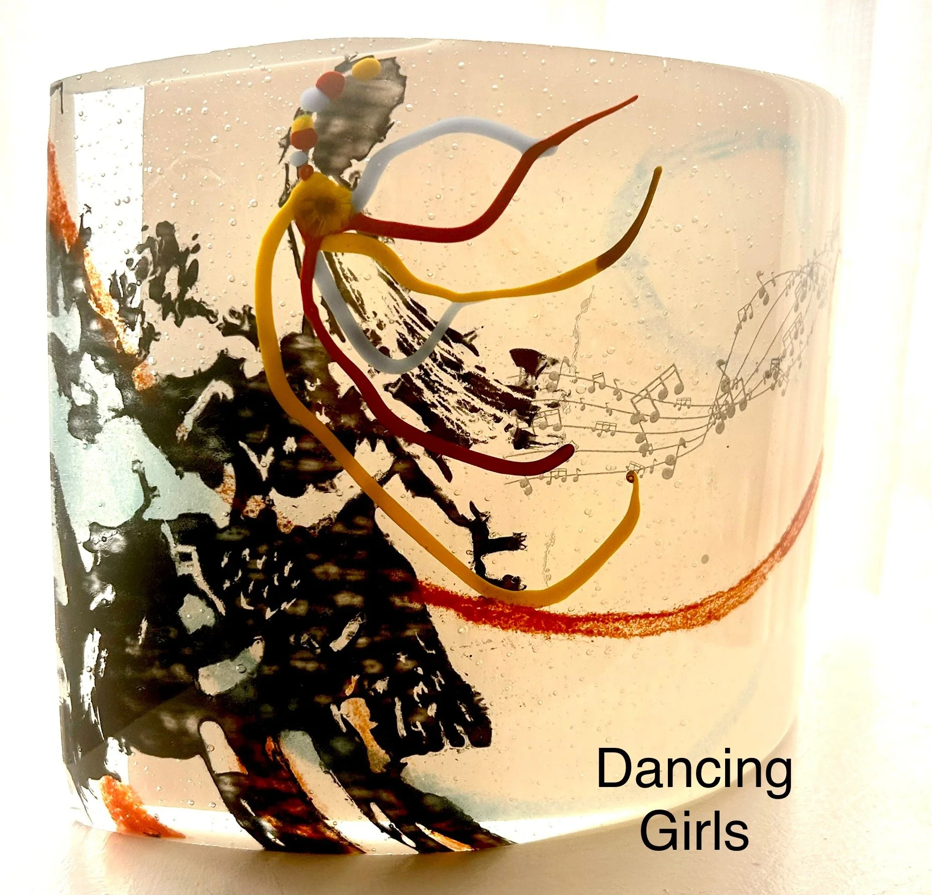 A glass with black and white artistic design, colorful wire decorations resembling girls dancing, musical notes, and the words "Dancing Girls" at the bottom right.