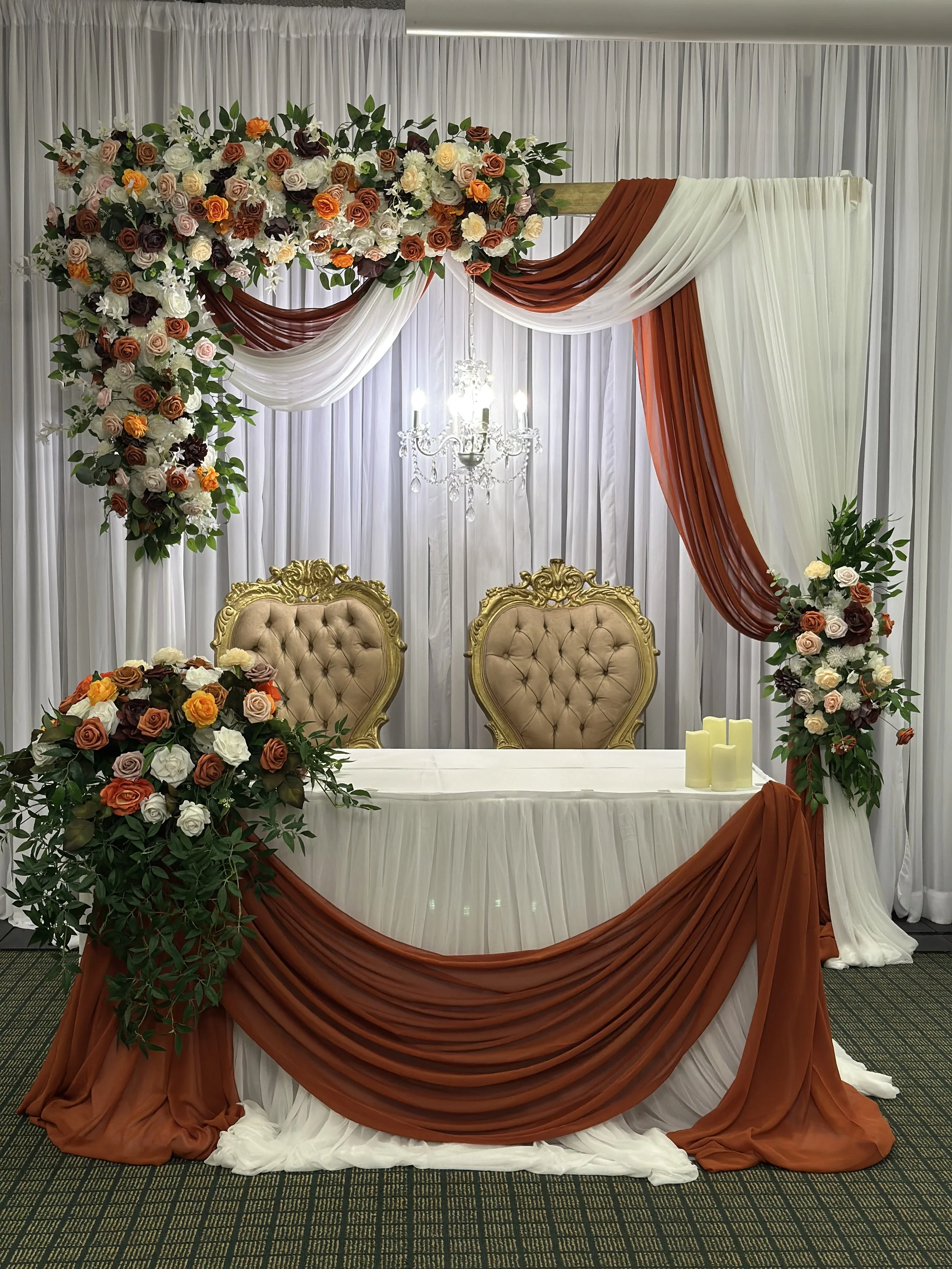 A wedding or event backdrop featuring white, rust, and dark red draped fabric with floral arrangements of roses in shades of white, peach, and deep red, with green foliage, over a white curtain backdrop. A table with a white tablecloth and candles in the foreground, along with two gold-colored upholstered chairs behind the table. A chandelier hangs above.