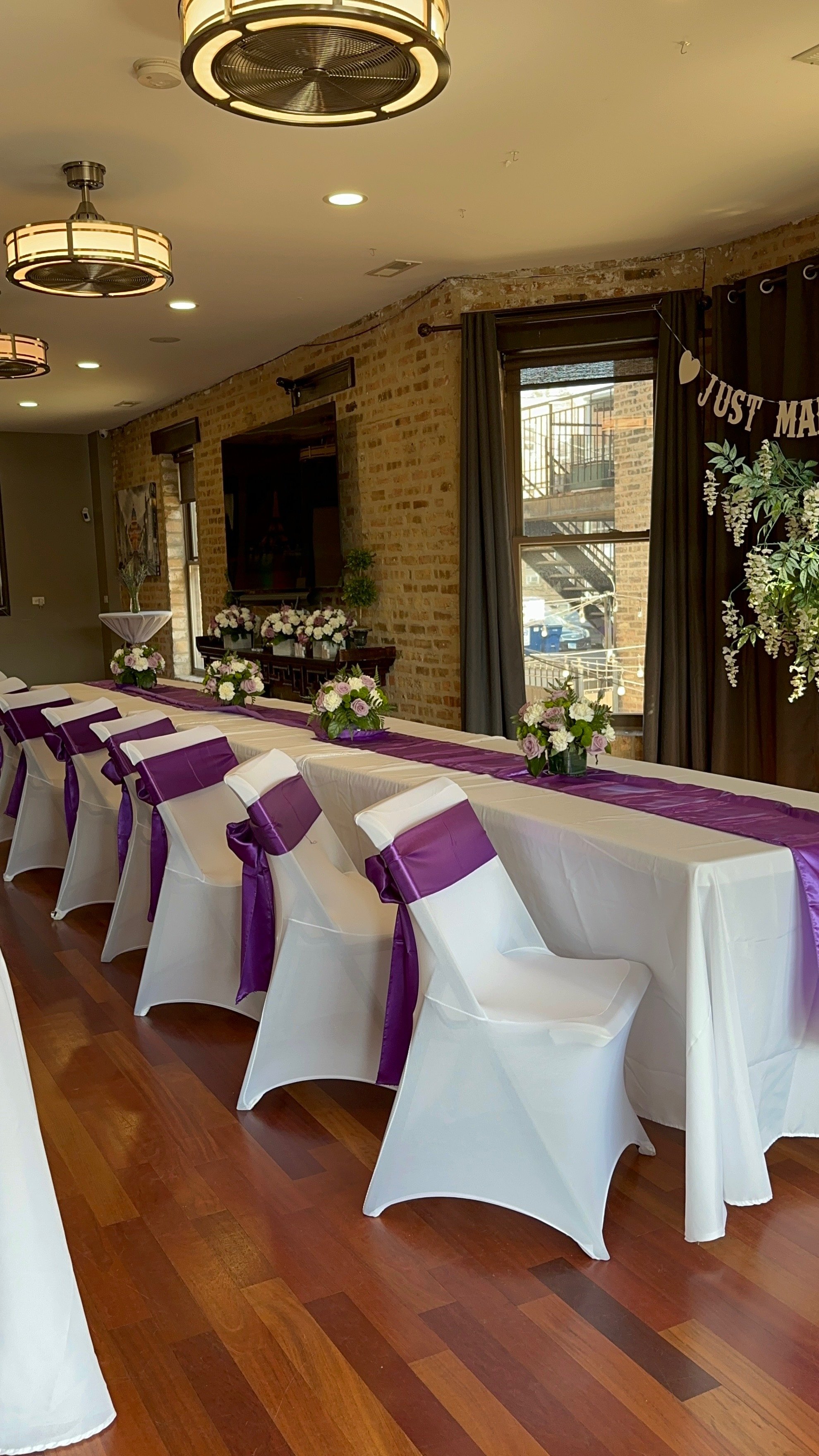 A decorated banquet hall for a wedding reception with a long table covered in white tablecloth and purple accents, surrounded by white chairs with purple sashes, floral centerpieces, a brick wall, a large window with dark curtains, and a