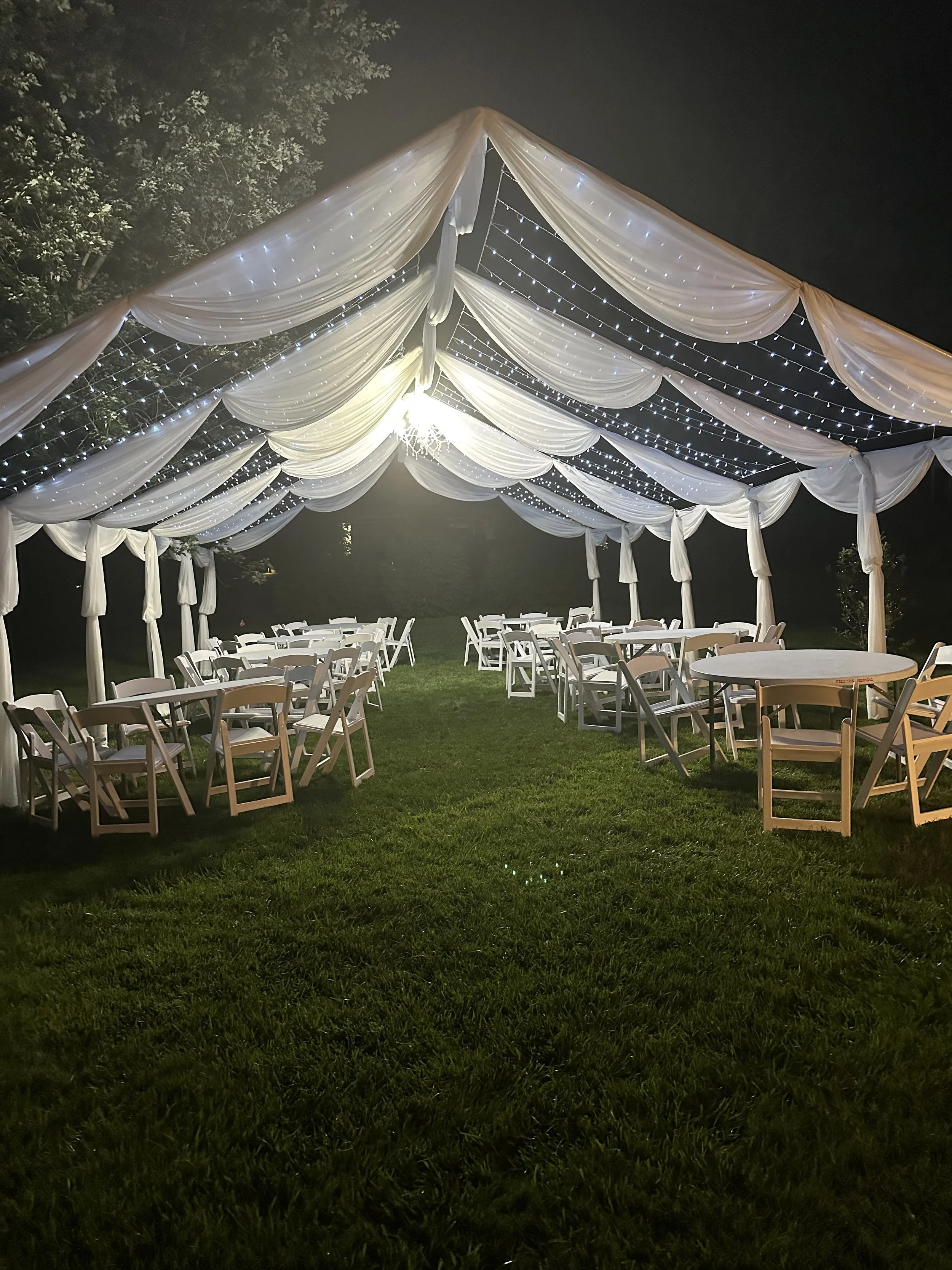 Outdoor event tent decorated with draped white fabric and string lights, with round tables and chairs on a grassy area at night.