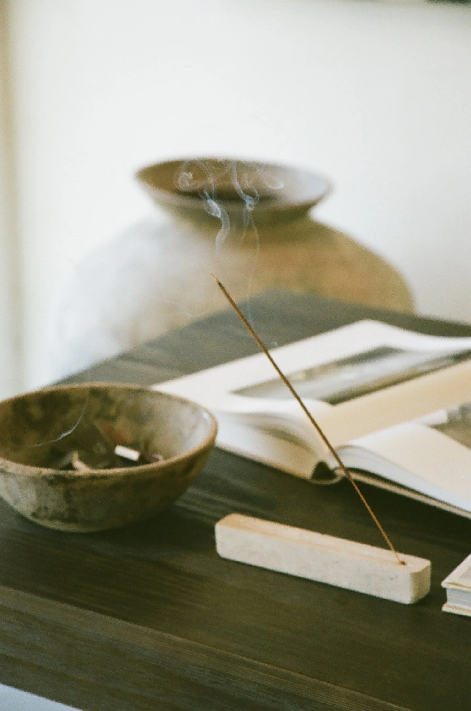 A wooden desk with a ceramic bowl, an open book, and a white base with a thin stick leaning on it, possibly an incense holder or decorative item.