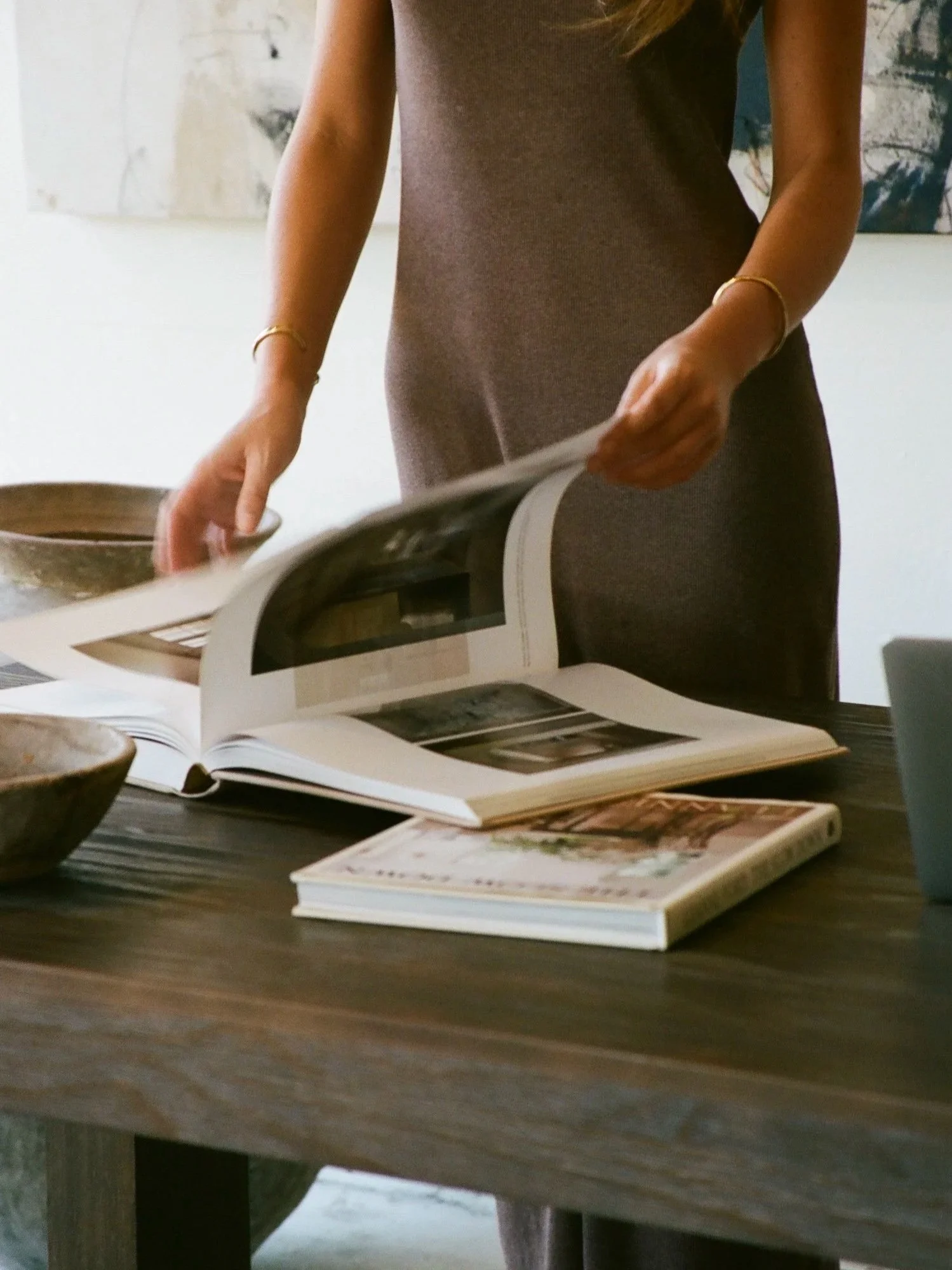A person with long hair wearing a brown dress is flipping through a magazine at a wooden table. There are additional magazines and bowls on the table.
