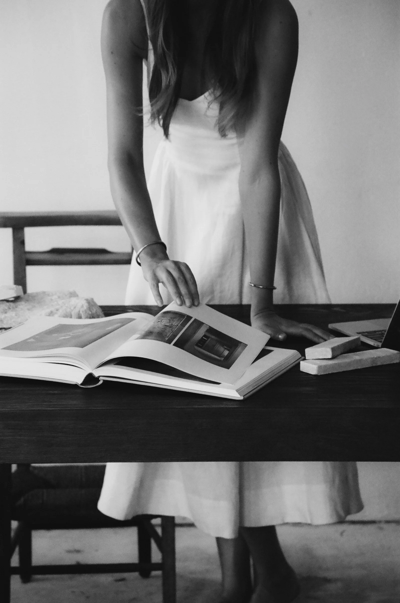 Black and white photograph of a woman in a sleeveless dress with long hair, partially visible, standing at a table with open books or magazines, her right hand resting on the table. The background includes a chair and plain wall.