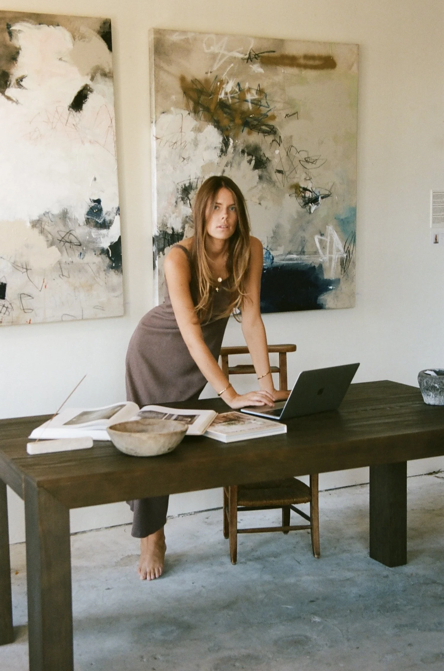A woman with long brown hair wearing a sleeveless brown top and pants is standing barefoot at a dark wooden table in an art gallery. She rests her hands on a laptop. The table has an open book and a bowl. Behind her are two abstract paintings with splashes of white, black, and brown, hung on white walls, with a white pillar separating them.