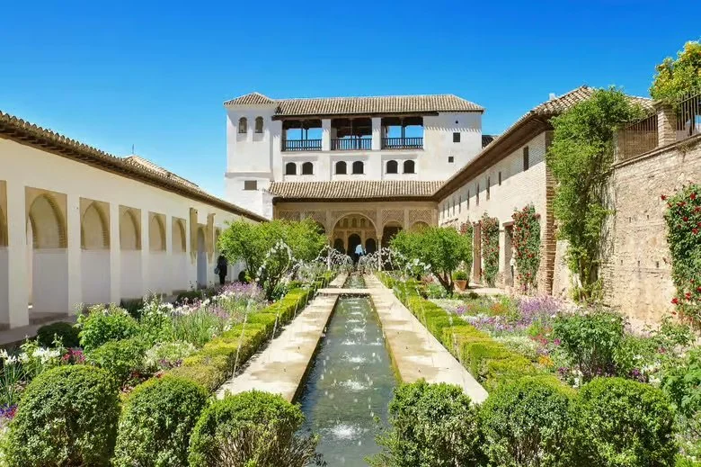 Jardín con fuente central en un palacio clásico, rodeado de muros con plantas y flores, en un día soleado y cielo azul.