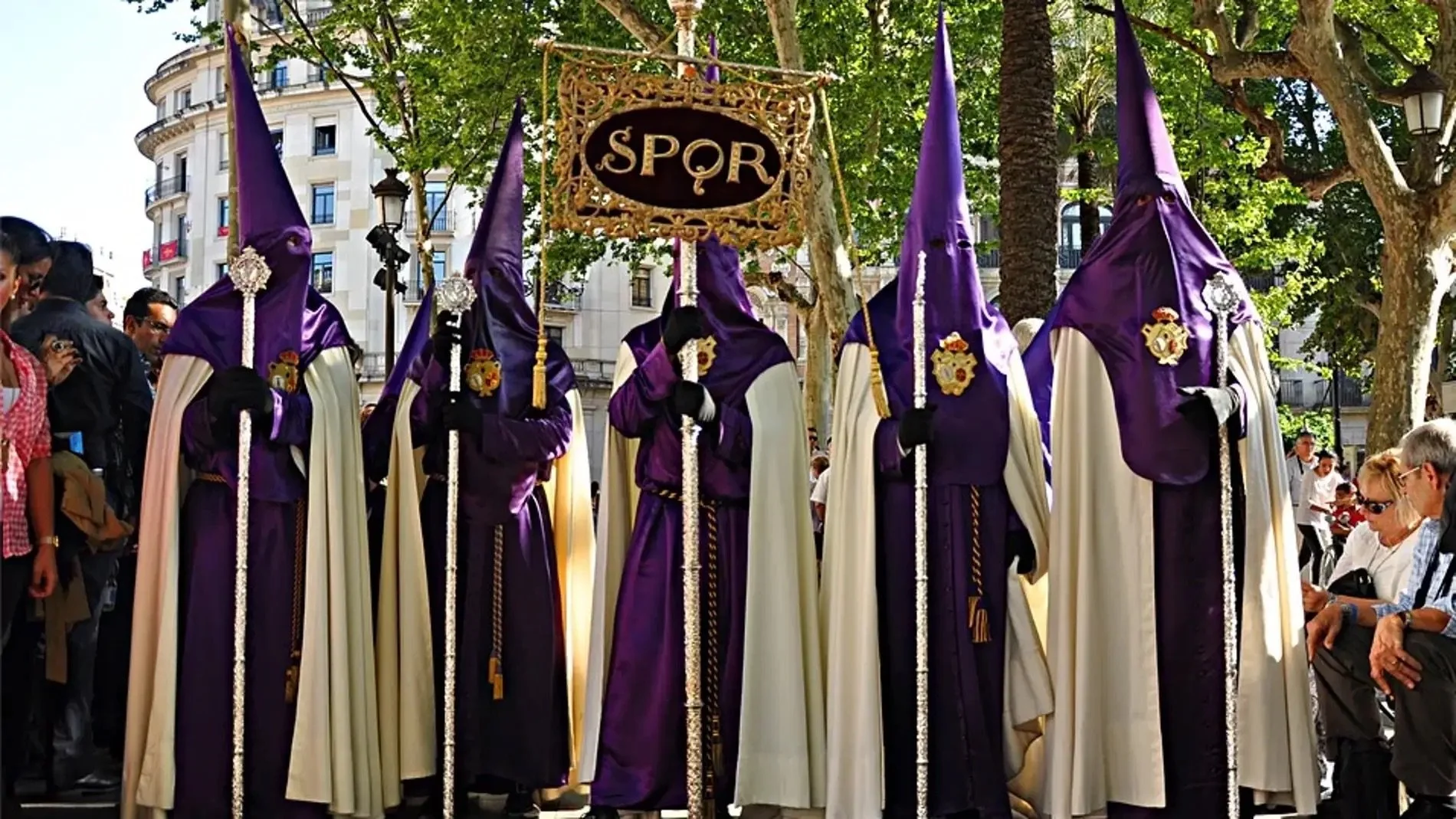 Nazarenos vestidos con túnicas moradas y capirotes participan en una procesión de la Semana Santa de Sevilla mientras sostienen cirios y el estandarte de su hermandad. La escena tiene lugar en una avenida del centro de la ciudad rodeada de árboles y 