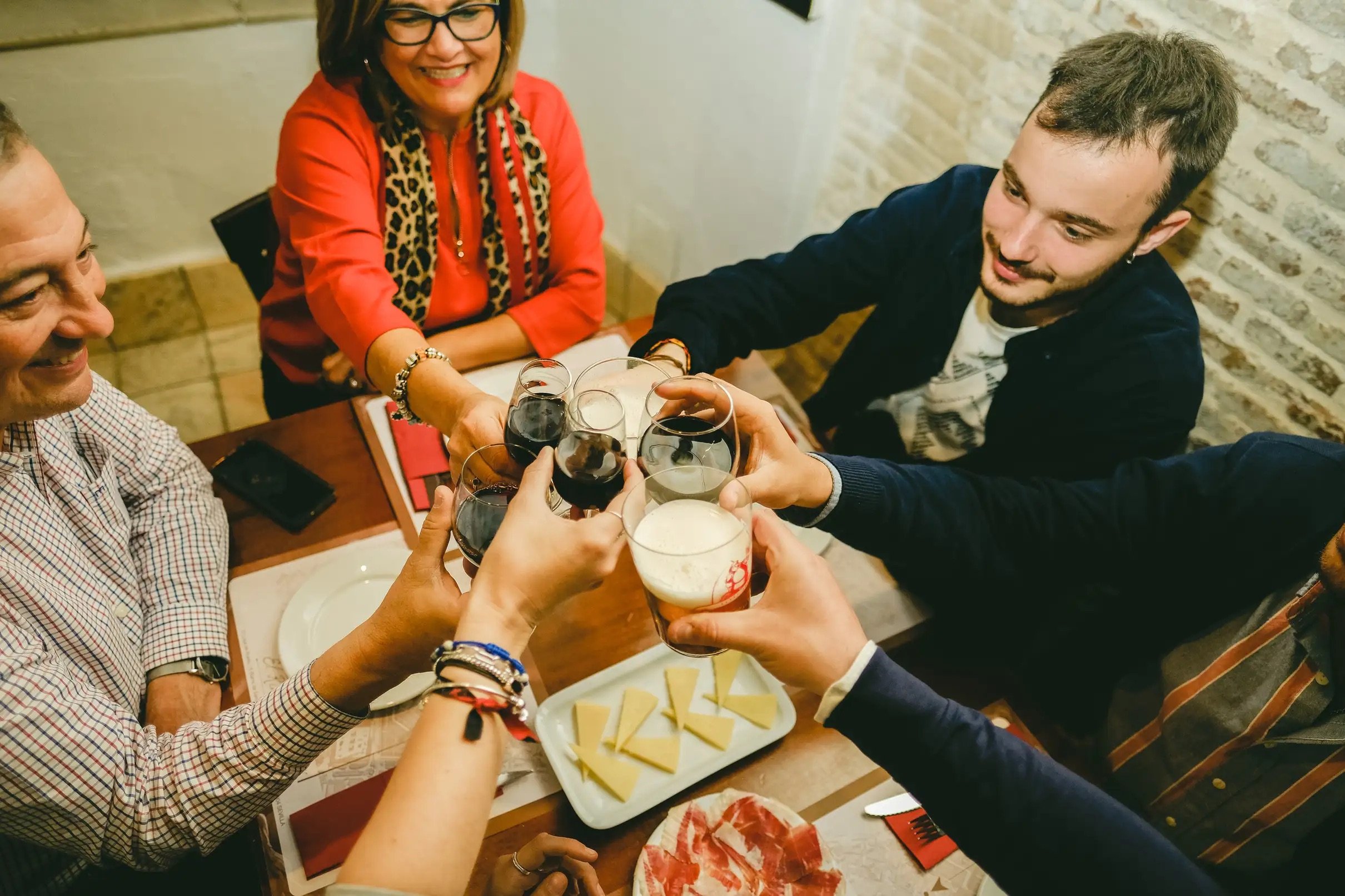 Grupo brindando con vino y cerveza durante una ruta gastronómica en Sevilla, disfrutando de tapas tradicionales como jamón ibérico y queso en un bar típico del centro histórico.