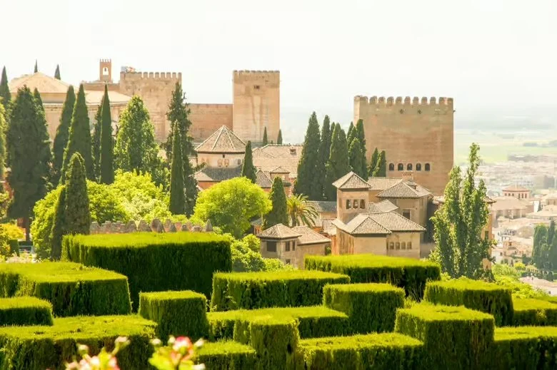 Vista del Alhambra de Granada con jardines verdes y arbustos recortados en primer plano, y árboles altos y edificios en el fondo, bajo cielo soleado.