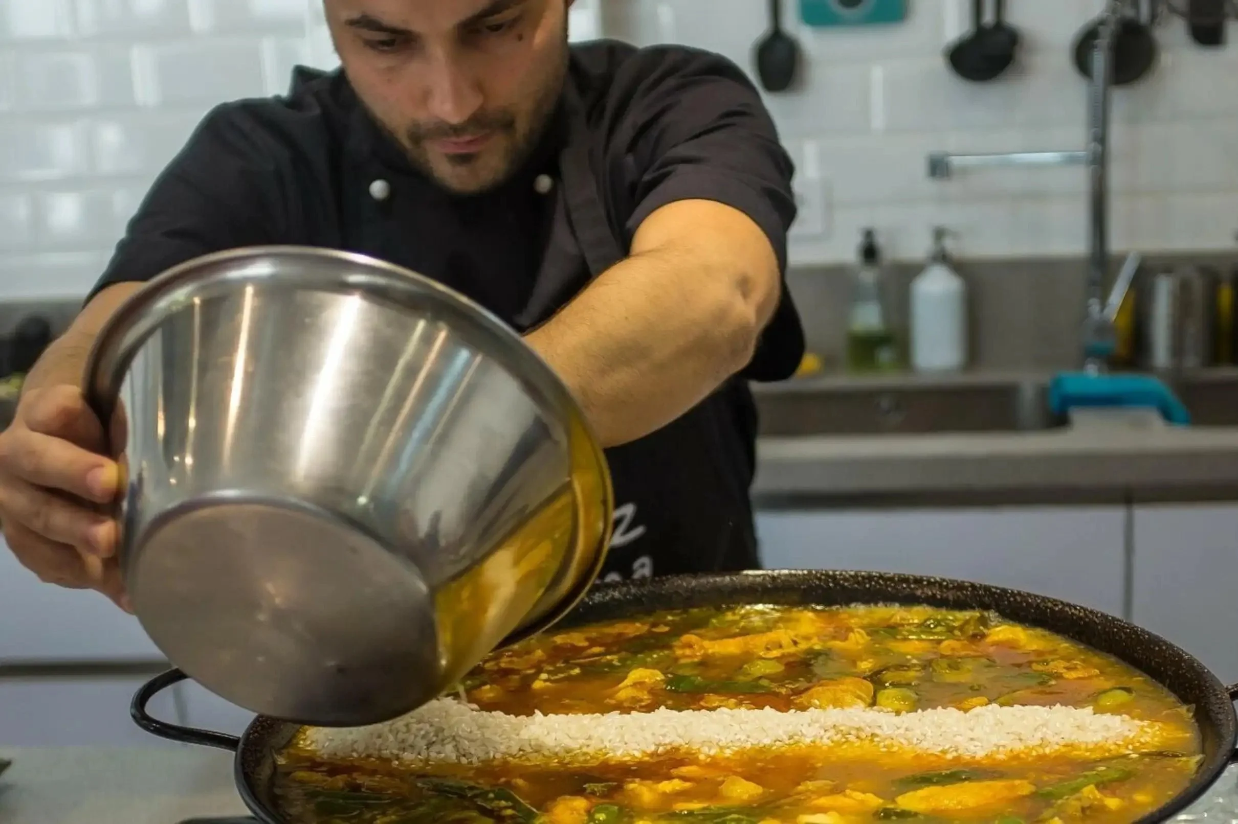 Chef preparando una paella durante una clase de cocina española en Sevilla, añadiendo arroz a la sartén en una experiencia gastronómica con cena incluida.