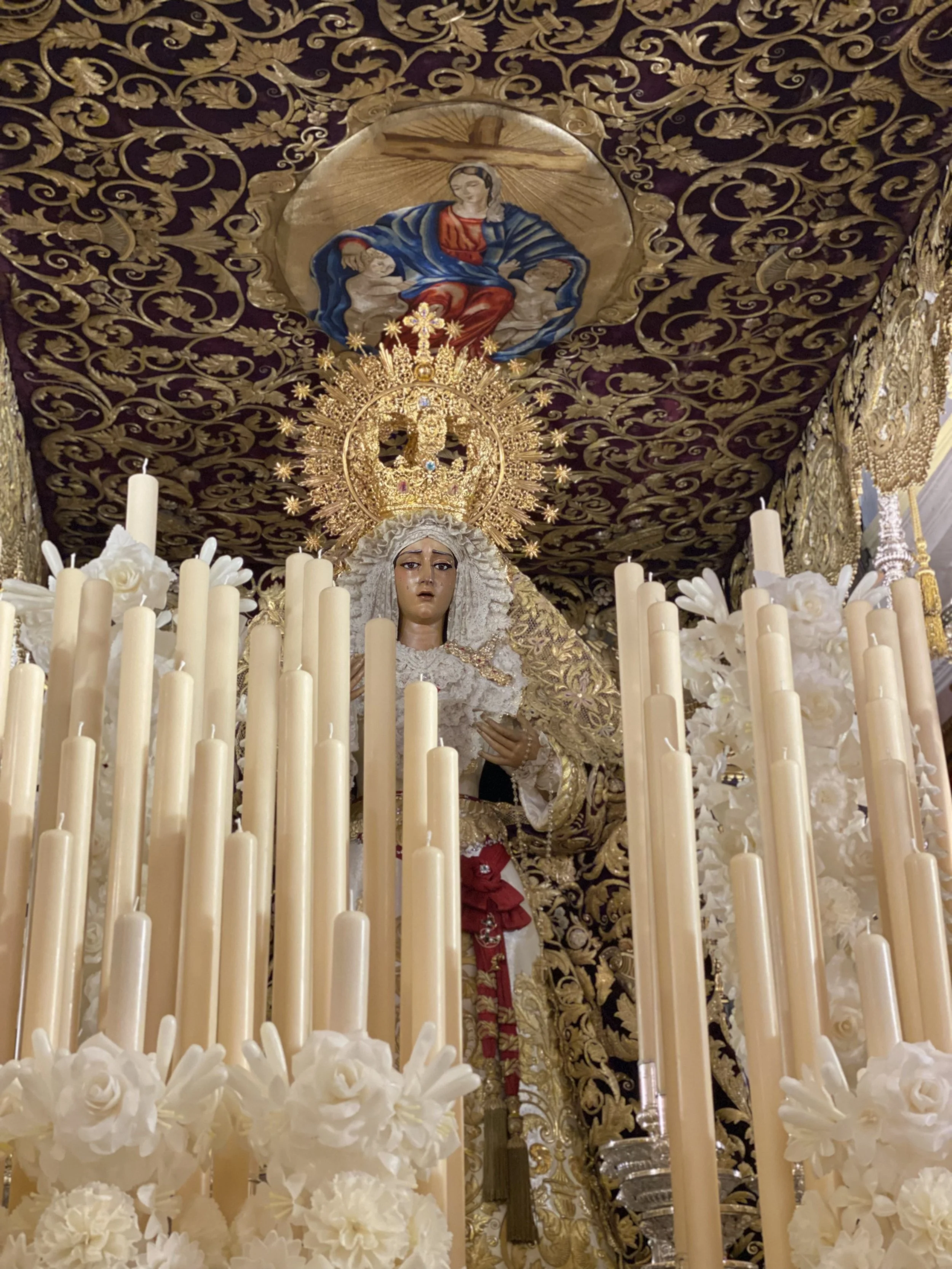 Virgen de la Piedad de la Hermandad del Baratillo, en paso de palio de la Semana Santa de Sevilla, rodeada de cirios encendidos y flores blancas, bajo un techo bordado que refleja la belleza, el arte y la devoción de esta tradición única.