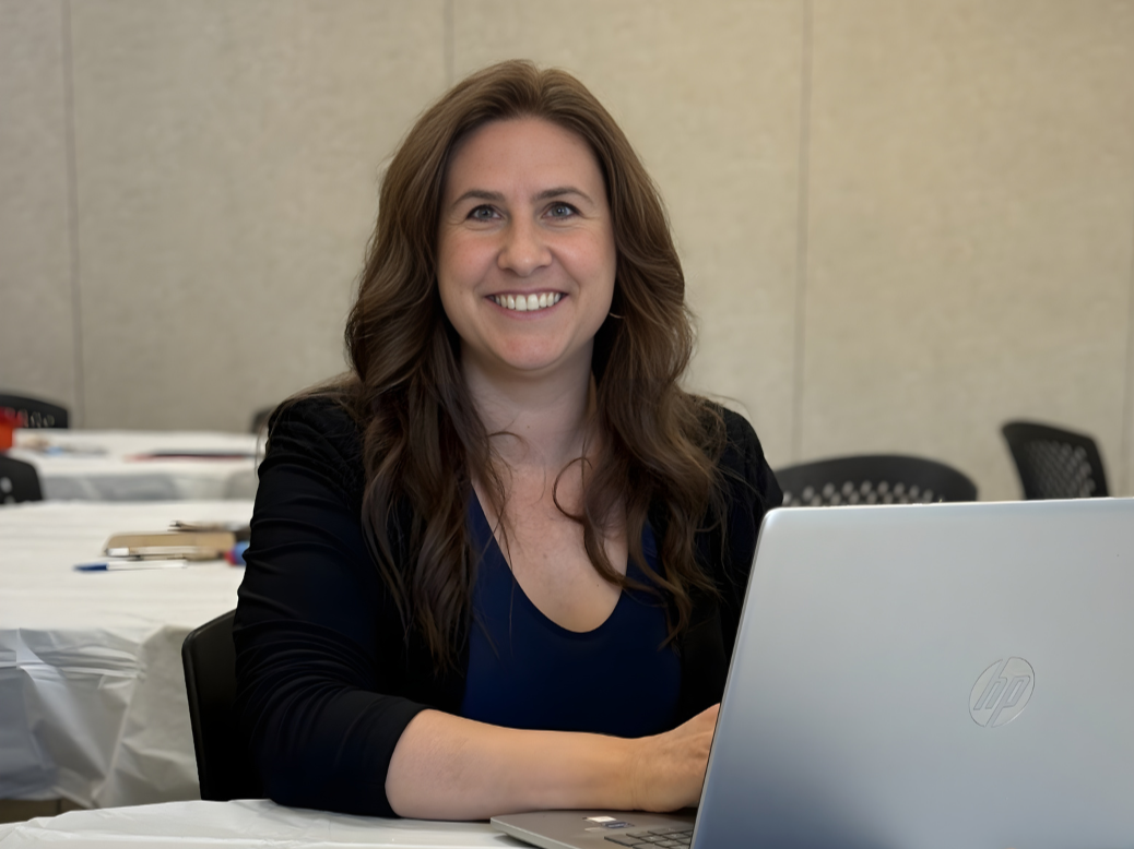 A woman with long brown hair sitting at a desk with a silver HP laptop, smiling at the camera in an office setting.