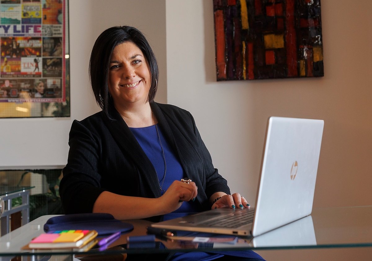 A woman with dark hair smiling at a laptop, seated at a glass desk with colorful stationery and small notebooks, in a room with modern art on the wall.