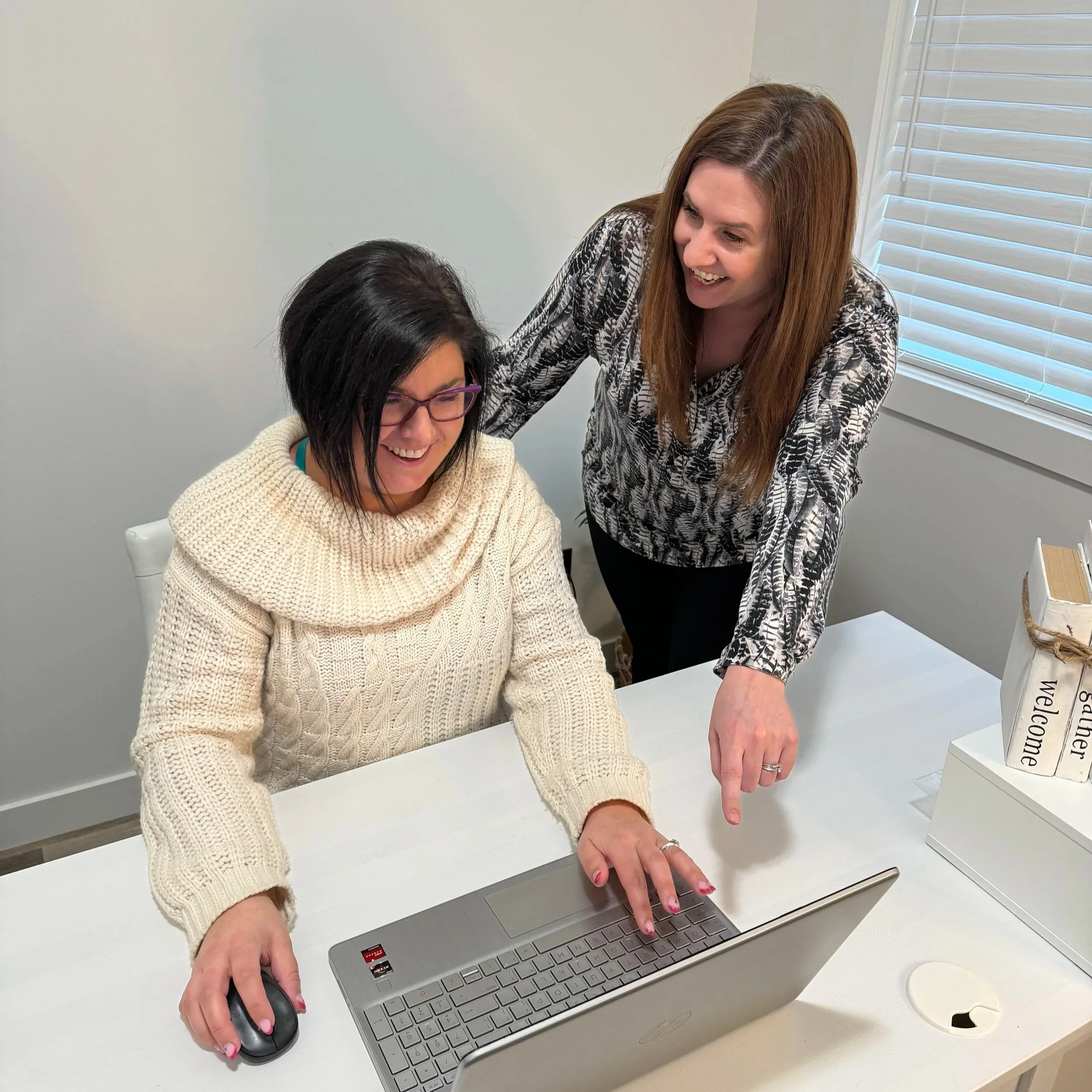 Two women working together at a desk, one seated using a laptop and mouse, the other standing and pointing at the laptop screen, smiling, in an office with a window and blind.