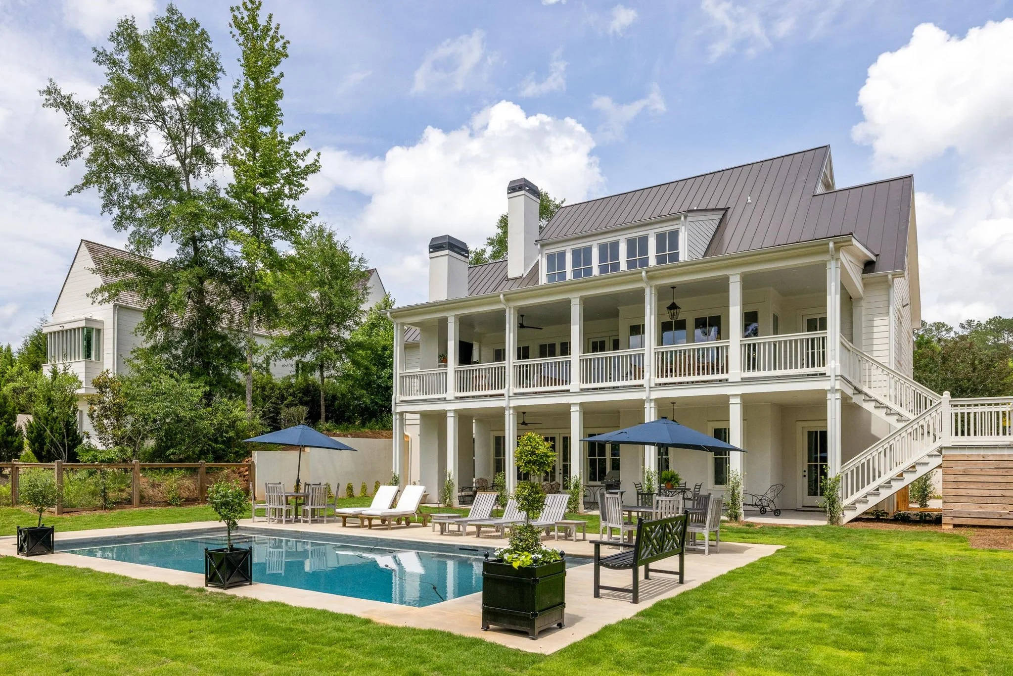 Southern-style home exterior with wraparound porch, blue umbrellas, and in-ground pool surrounded by manicured landscaping