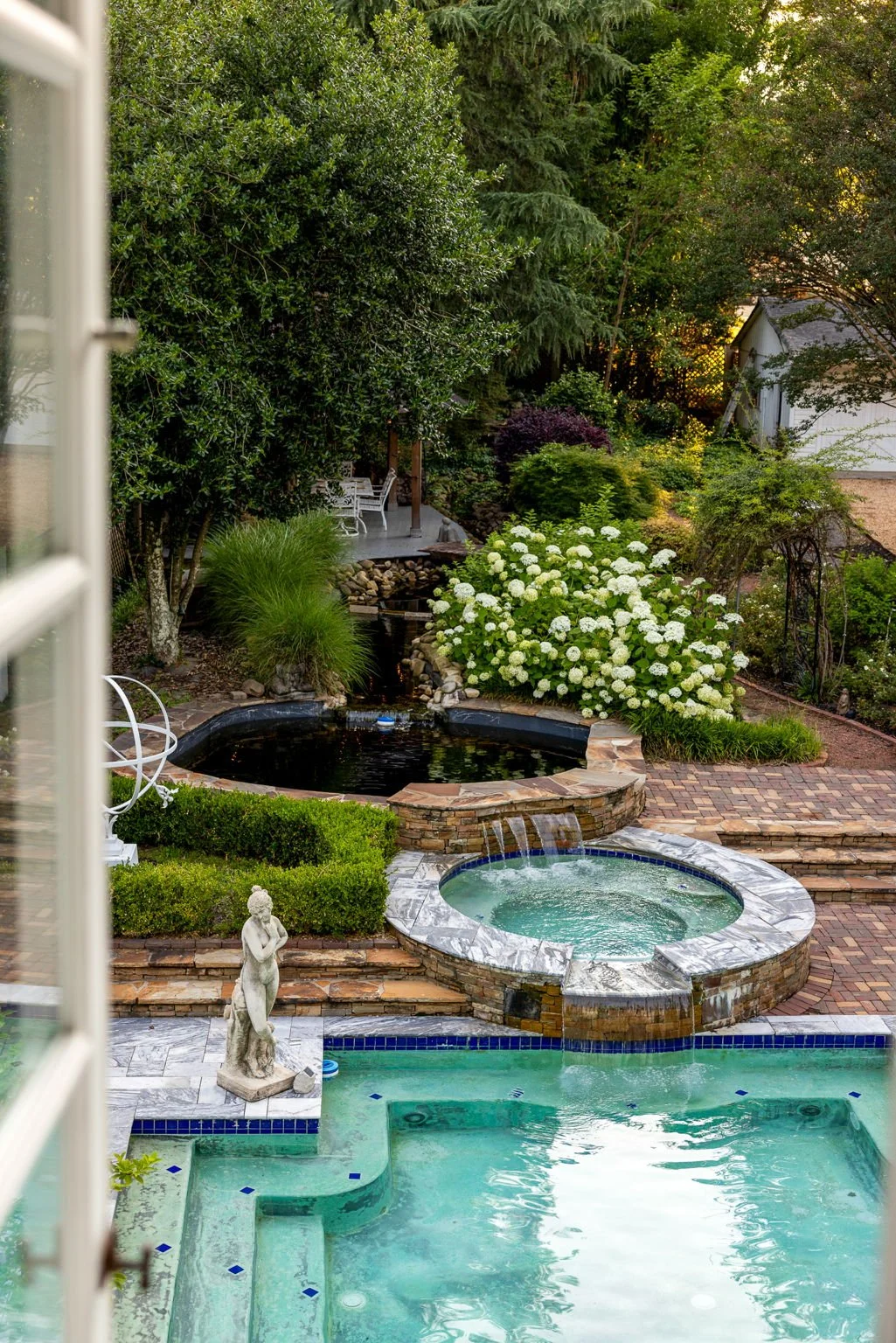 A view from a bedroom window out over a pool  and hot tub set in a landscaped garden.