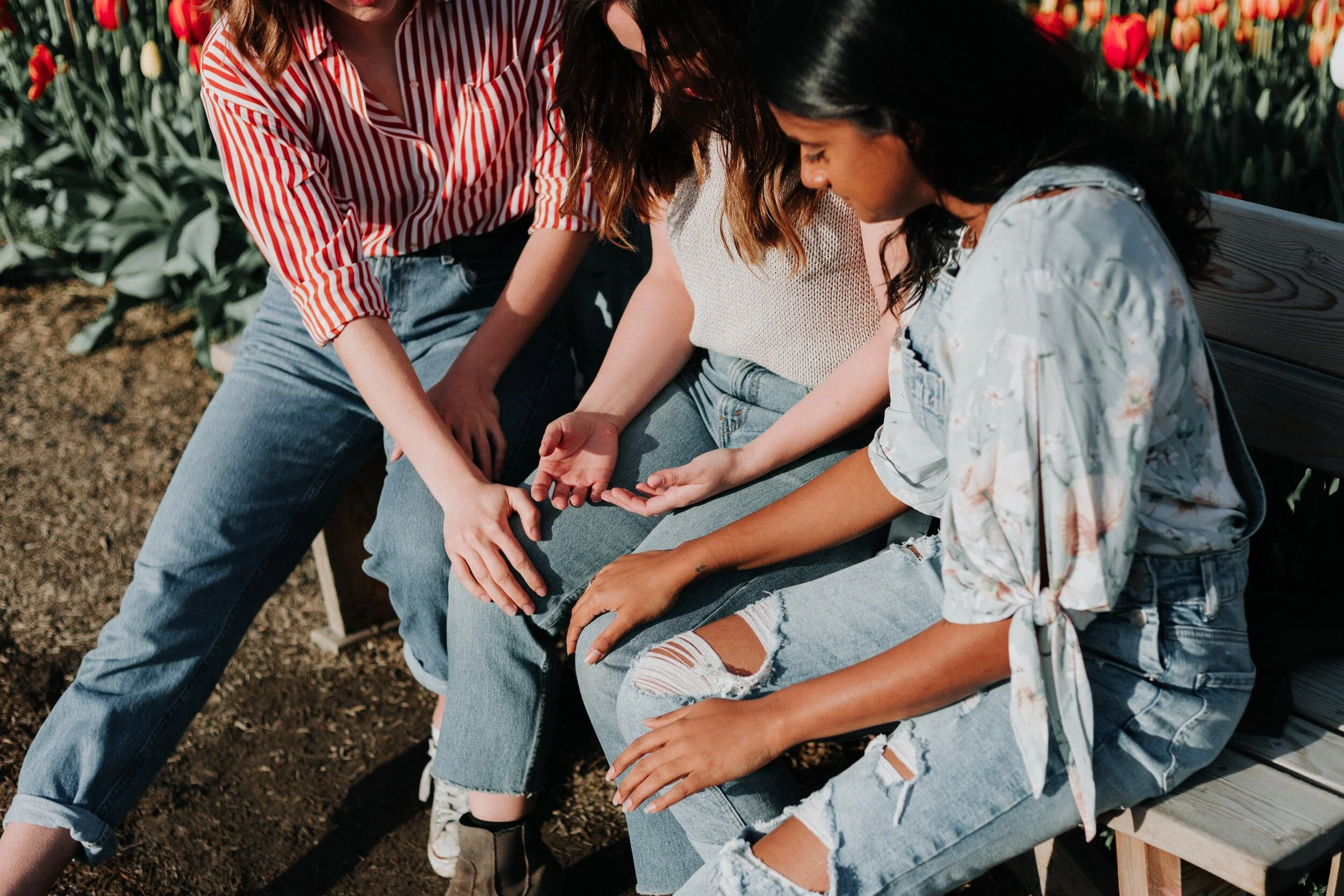 Three women sitting on a park bench, one woman gently touching the hand of another woman, in a garden with red and yellow tulips in the background.