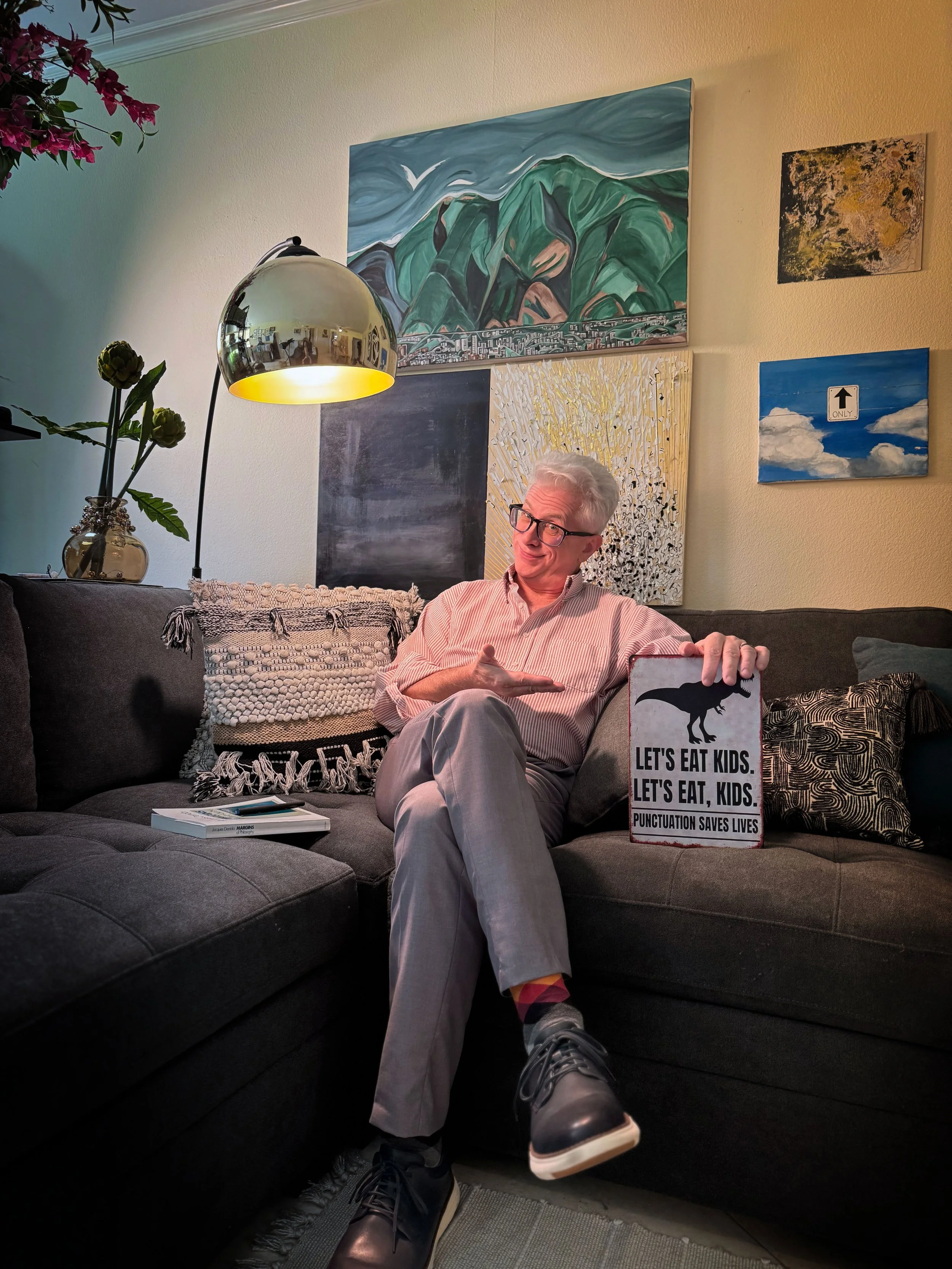 Anthony Krupp, is holding a sign that reads "Let's eat kids. Let's eat, kids. Punctuation saves lives," with a dinosaur silhouette. The room has a yellow wall decorated with various paintings and art, a tall lamp with a reflective metallic shade
