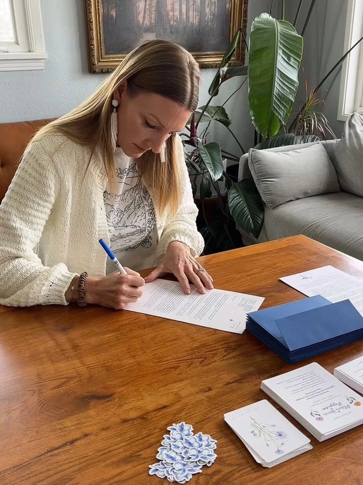 A woman sitting at a wooden table signing a document with a blue pen. She is wearing a white cardigan, floral shirt, and long earrings. On the table are brochures, pamphlets, and decorative floral cutouts. Behind her are houseplants, a grey couch, and framed artwork on the wall.