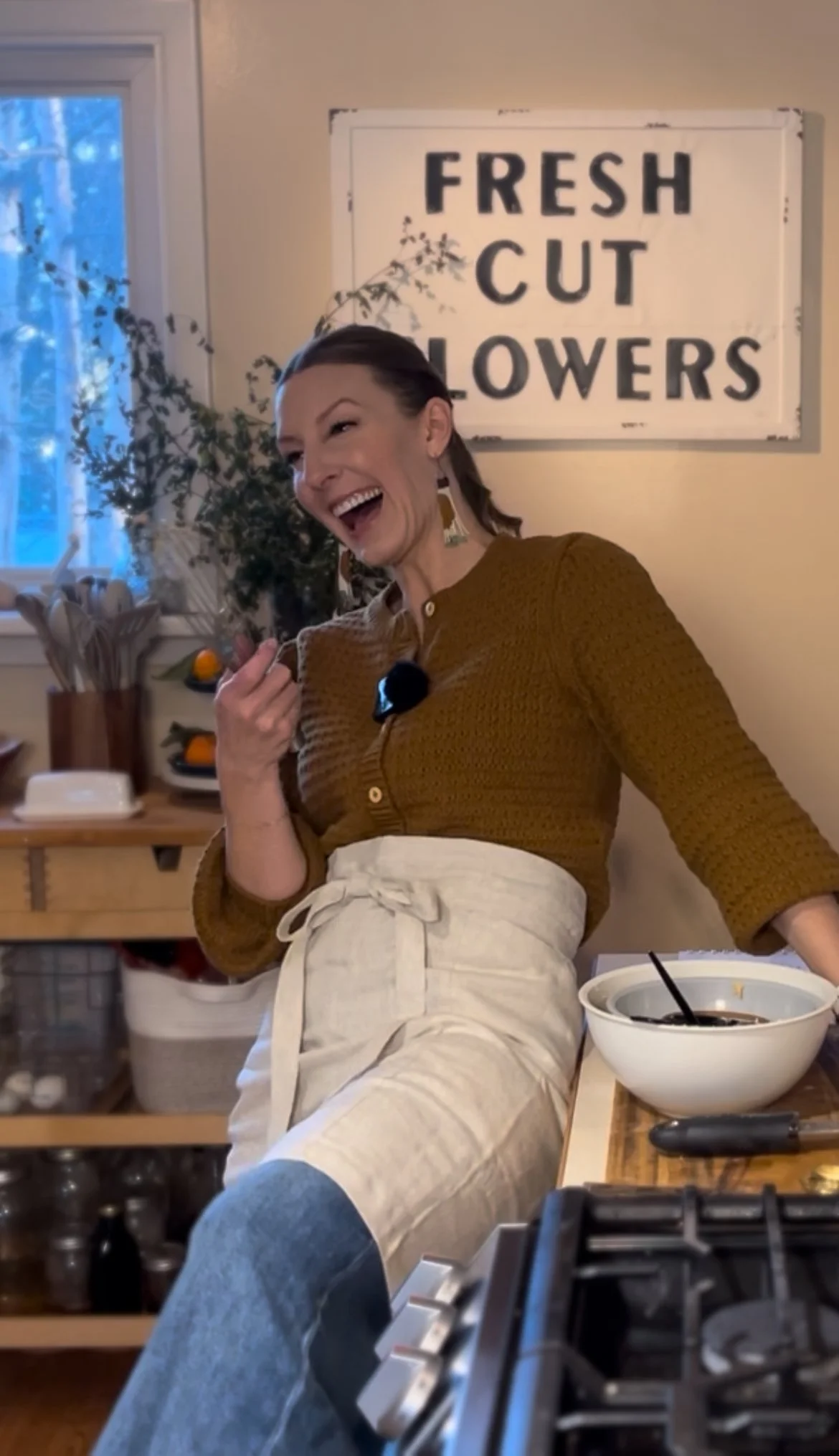 A woman laughing in a kitchen, holding a small flower bouquet, standing in front of a sign that reads "FRESH CUT FLOWERS." She is wearing a brown top and beige apron, with a bowl of food and a spoon nearby.