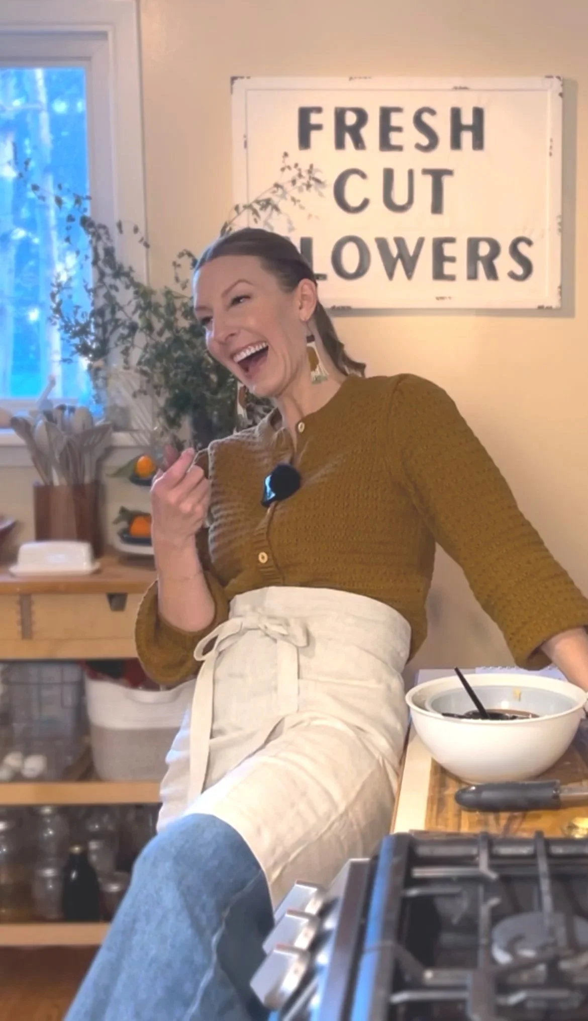 A woman in a mustard yellow sweater and beige apron leaning on a kitchen counter, smiling and laughing, with a sign that reads 'FRESH CUT FLOWERS' on the wall behind her. There are plants and kitchen items in the background.