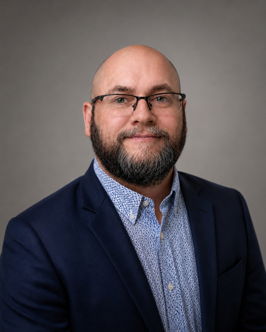 A headshot of a man with a bald head, glasses, and a beard, wearing a navy blue suit and a patterned light blue shirt, against a gray background.