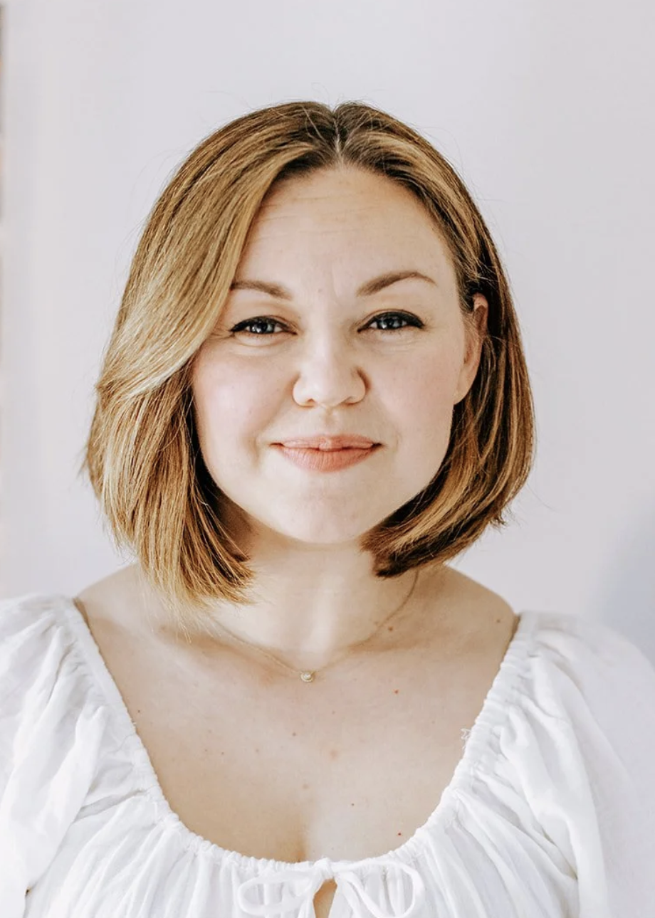 A woman with shoulder-length reddish-brown hair, smiling, wearing a white gathered blouse with a bow at the neckline.