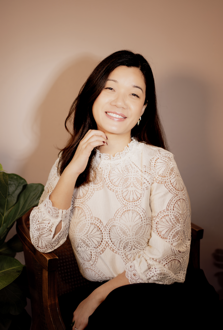 A woman with long dark hair smiling and sitting on a wooden chair near a green leafy plant, wearing a white lace top and earrings, against a neutral beige background.