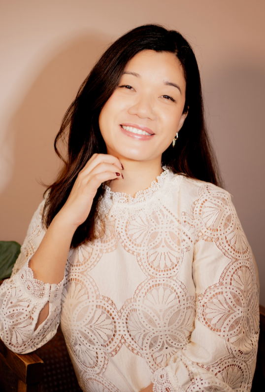 A woman with dark hair and earrings, smiling, wearing a navy blue top, indoors with a blurred background.