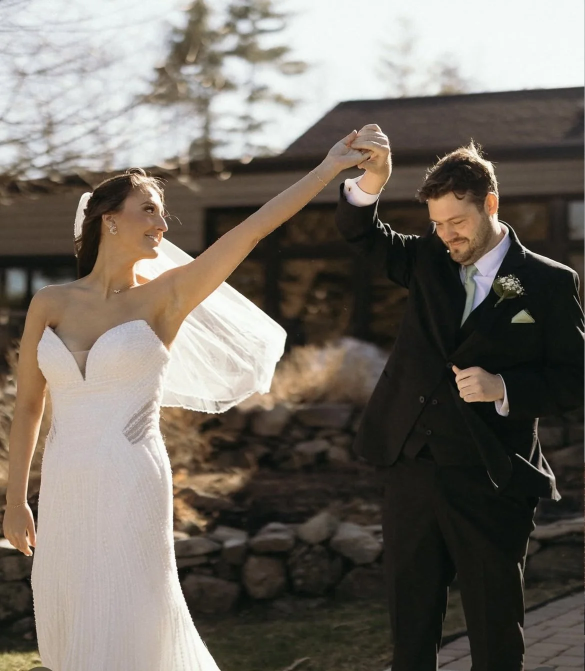 A bride and groom dancing outdoors on a sunny day, with the bride wearing a white strapless wedding dress and veil, and the groom in a black suit, gently holding hands.