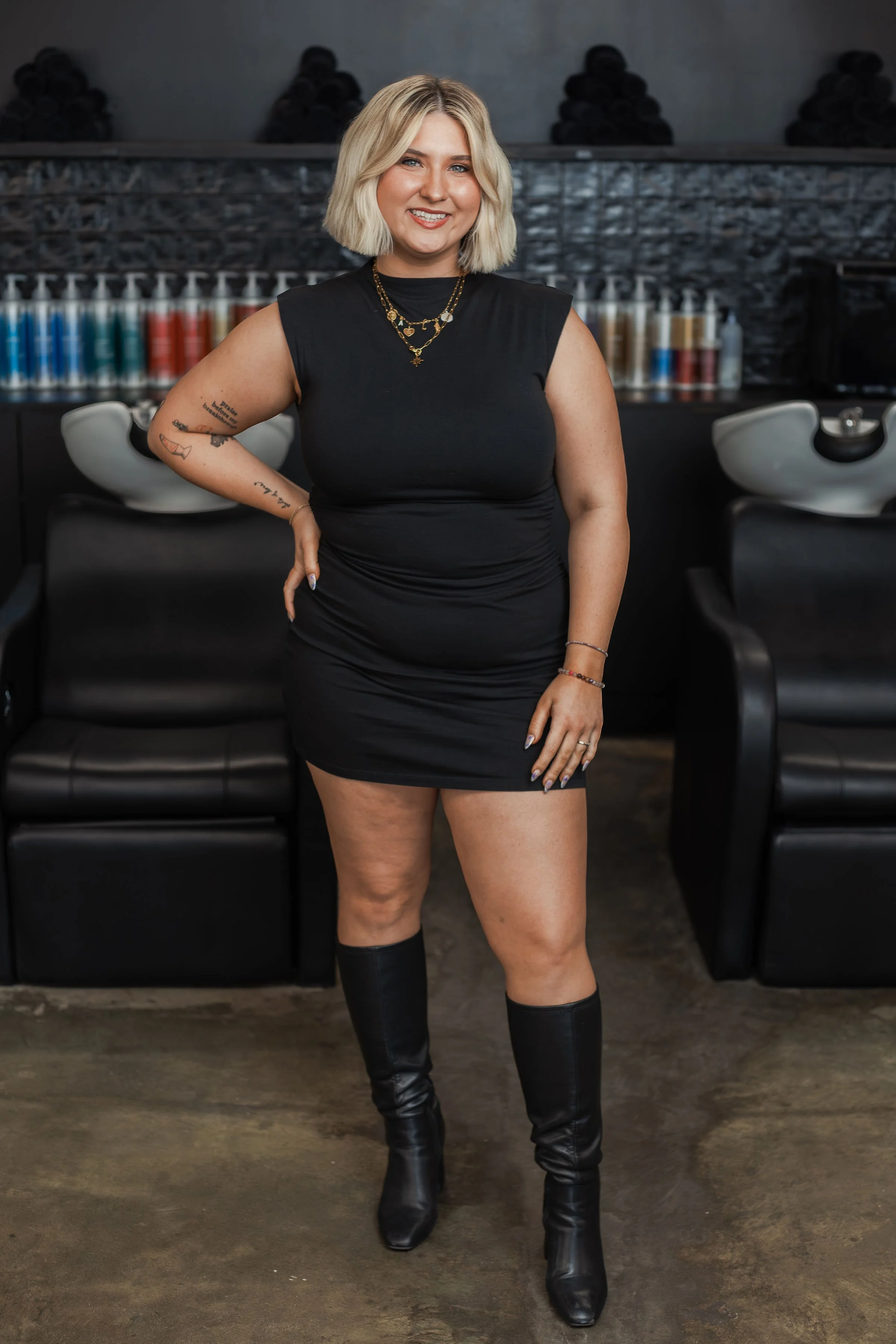 A woman with blonde, shoulder-length hair smiling, dressed in a black sleeveless dress and black knee-high boots, standing in a salon or barbershop with salon chairs, hair products, and hair dryers in the background.